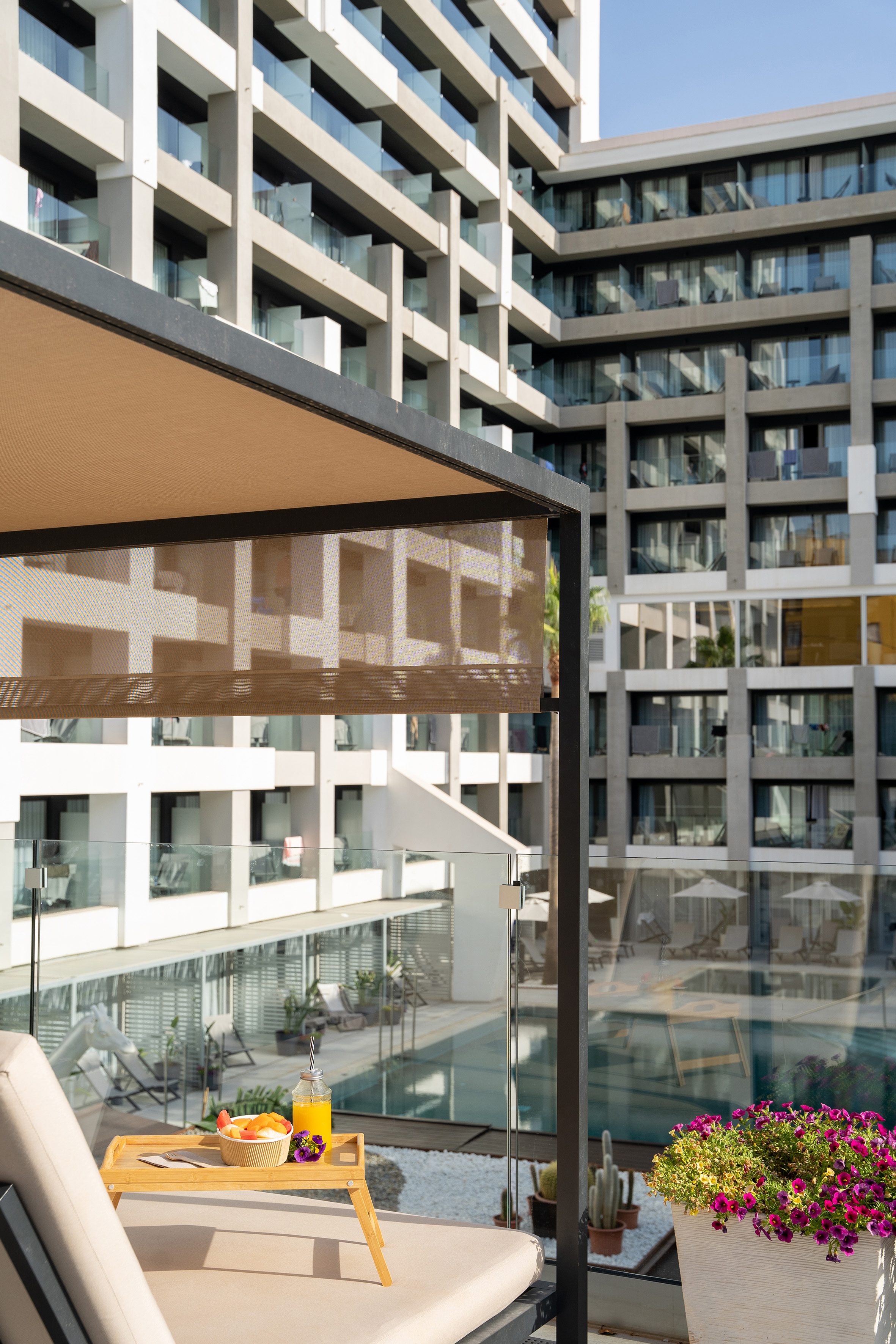 Hotel balcony breakfast: fruit, juice, modern resort building, pool view.