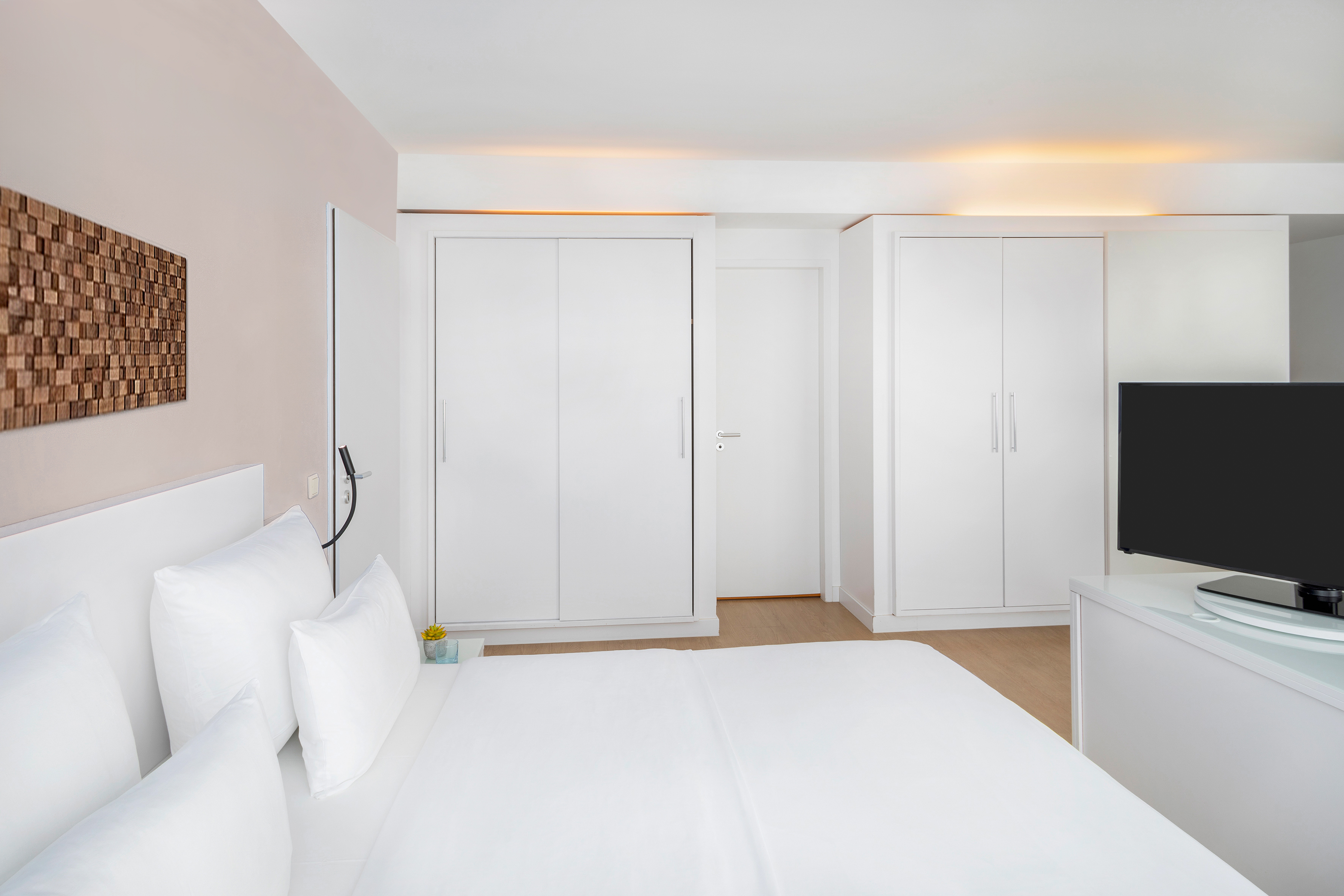 Modern white hotel bedroom with bed, built-in wardrobes, TV, wood floor.