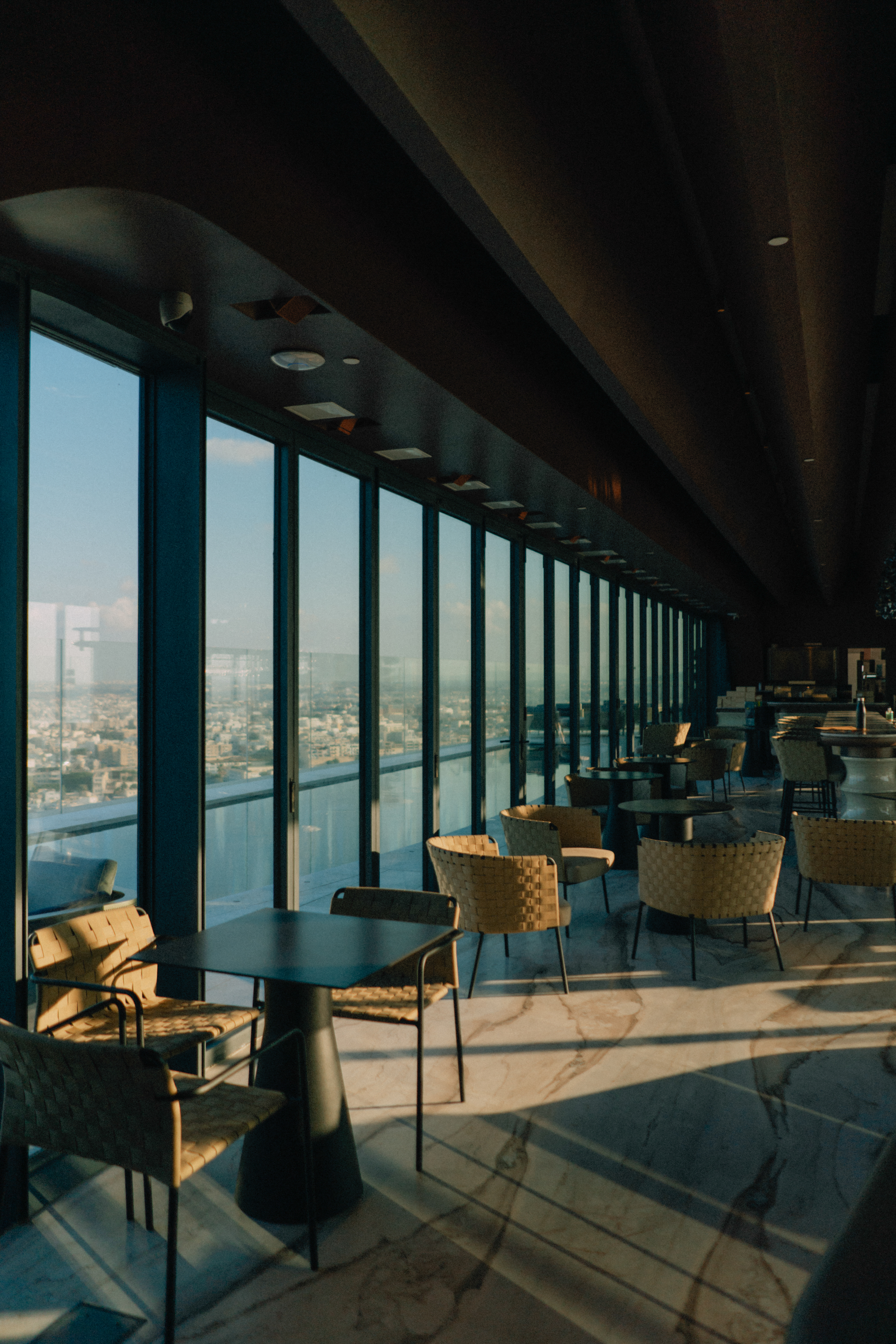 Upscale lounge with panoramic city views, modern tables, and woven chairs.