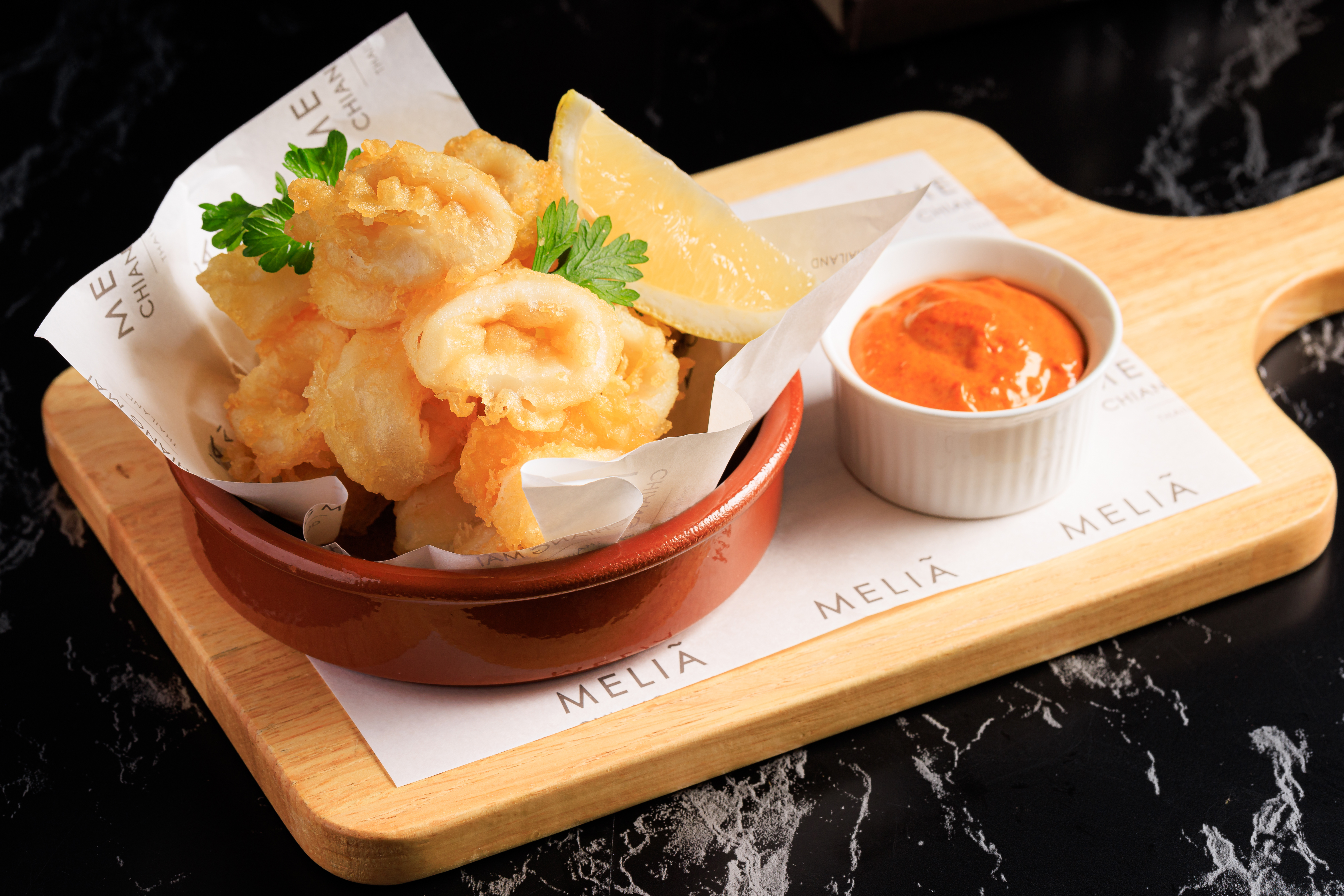 Crispy fried calamari, lemon, parsley, and red dipping sauce on Melia branded paper.