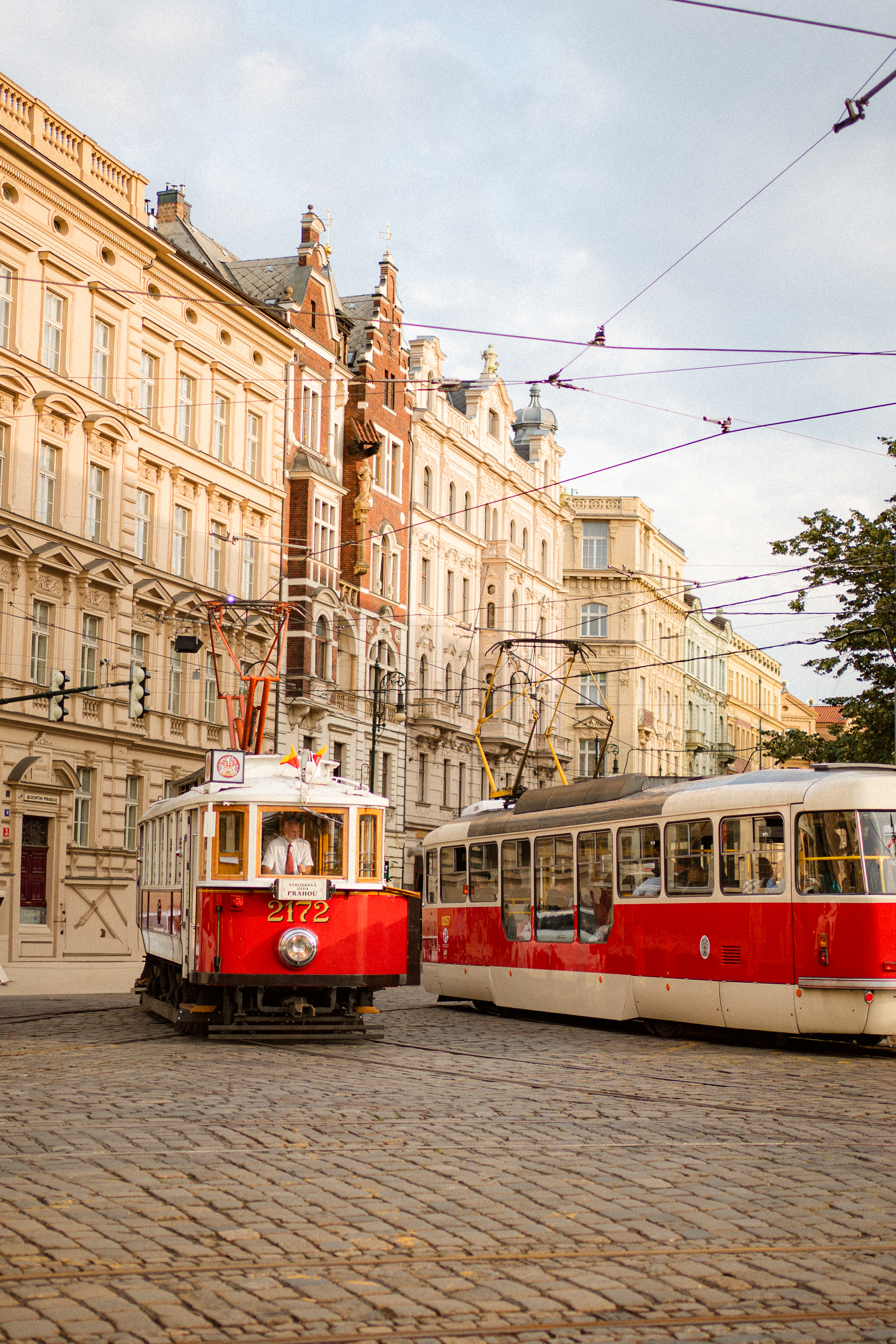 Red and white Prague trams on a cobblestone street with historic buildings.
