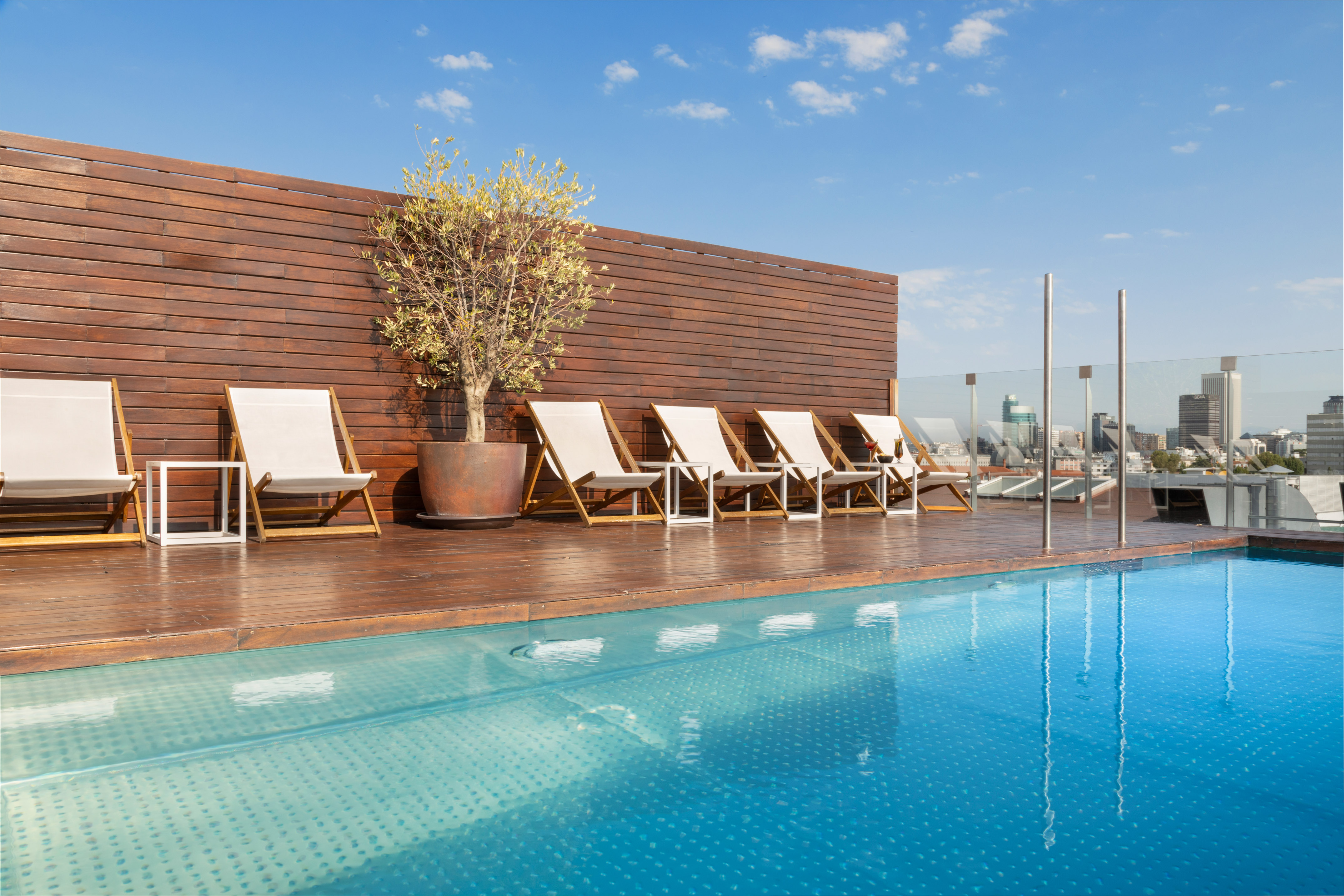 Rooftop swimming pool with white loungers, wood deck, and city skyline.