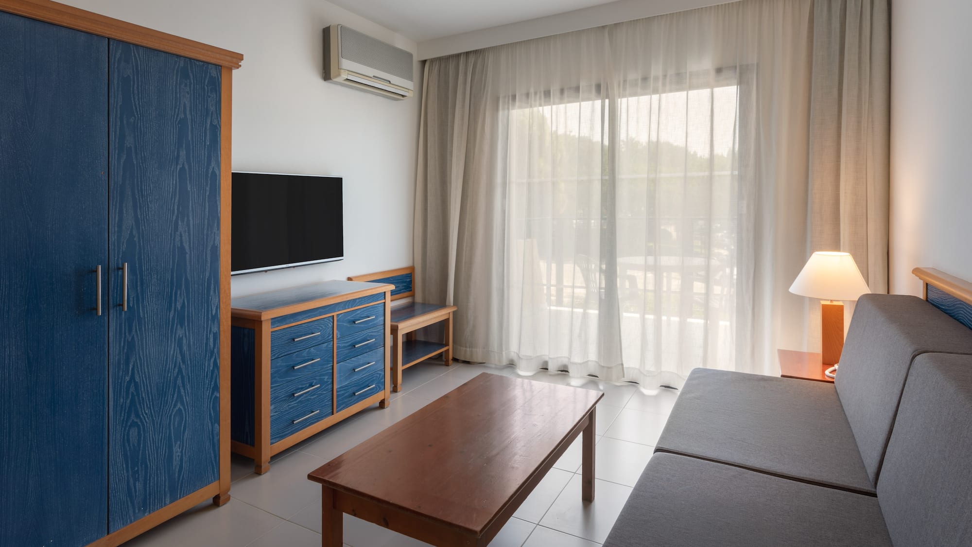 Modern hotel living room with blue cabinet, TV, gray sofa, and balcony view.