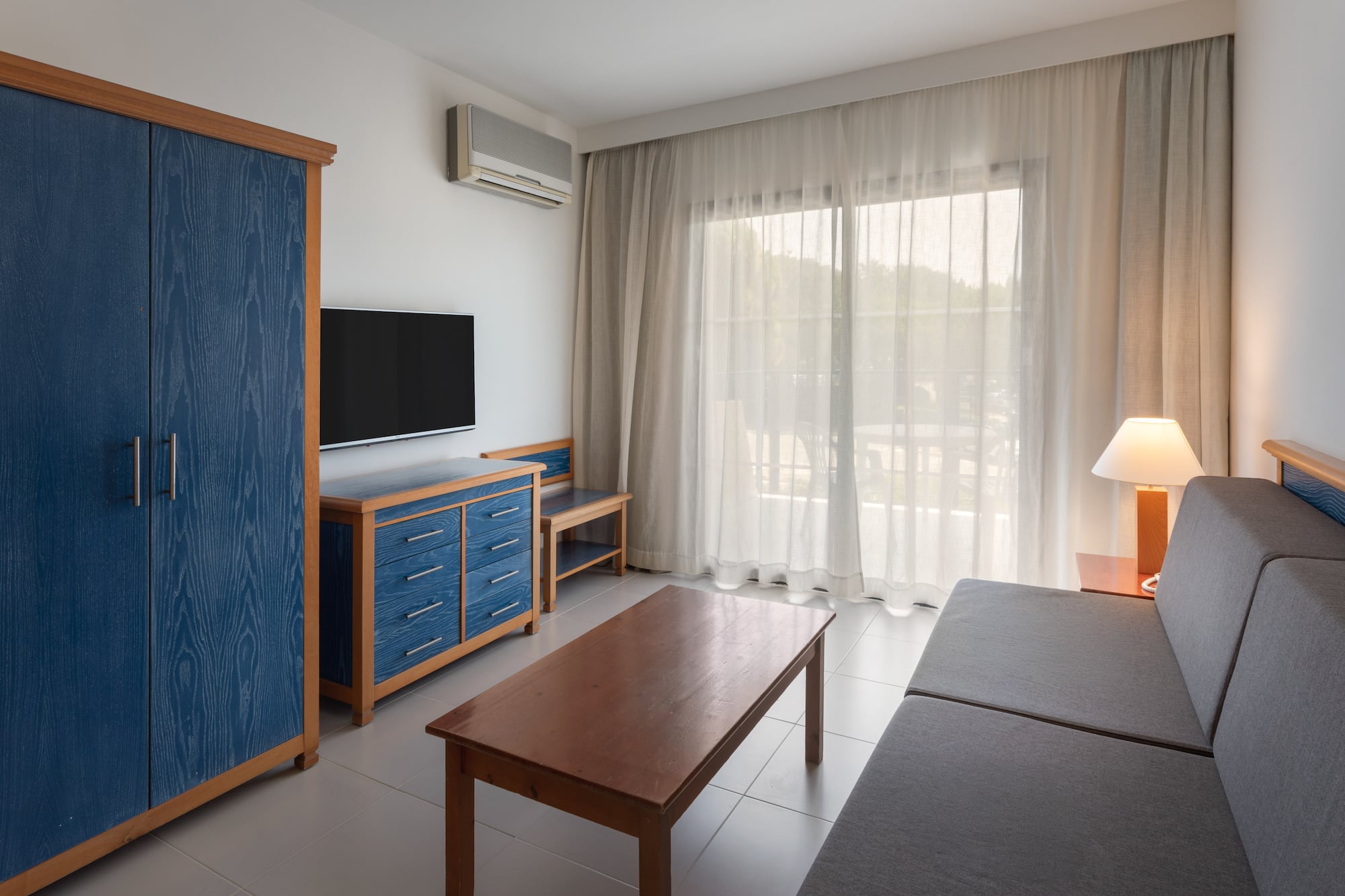 Modern hotel living room with blue cabinet, TV, gray sofa, and balcony view.