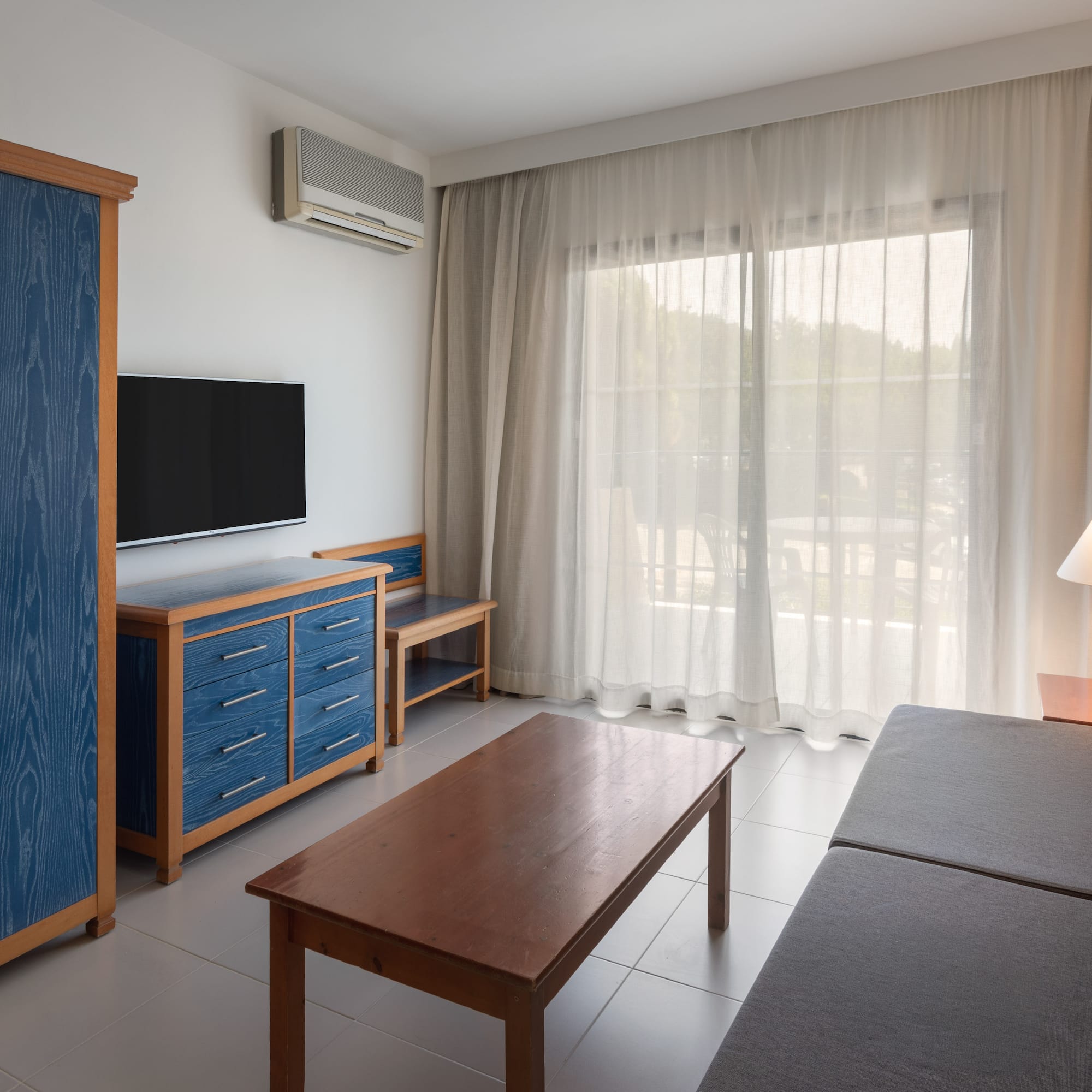 Modern hotel living room with blue cabinet, TV, gray sofa, and balcony view.