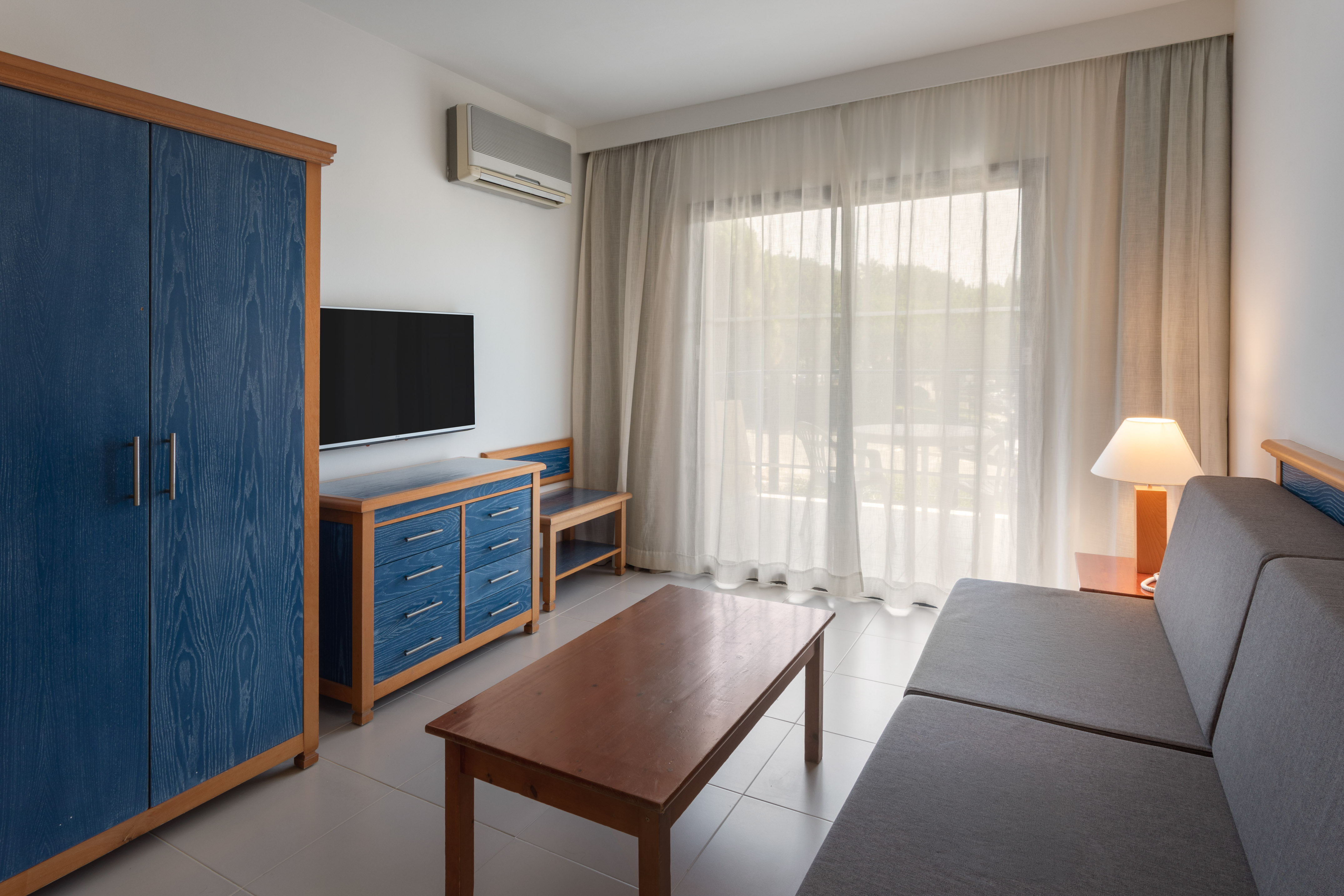 Modern hotel living room with blue cabinet, TV, gray sofa, and balcony view.