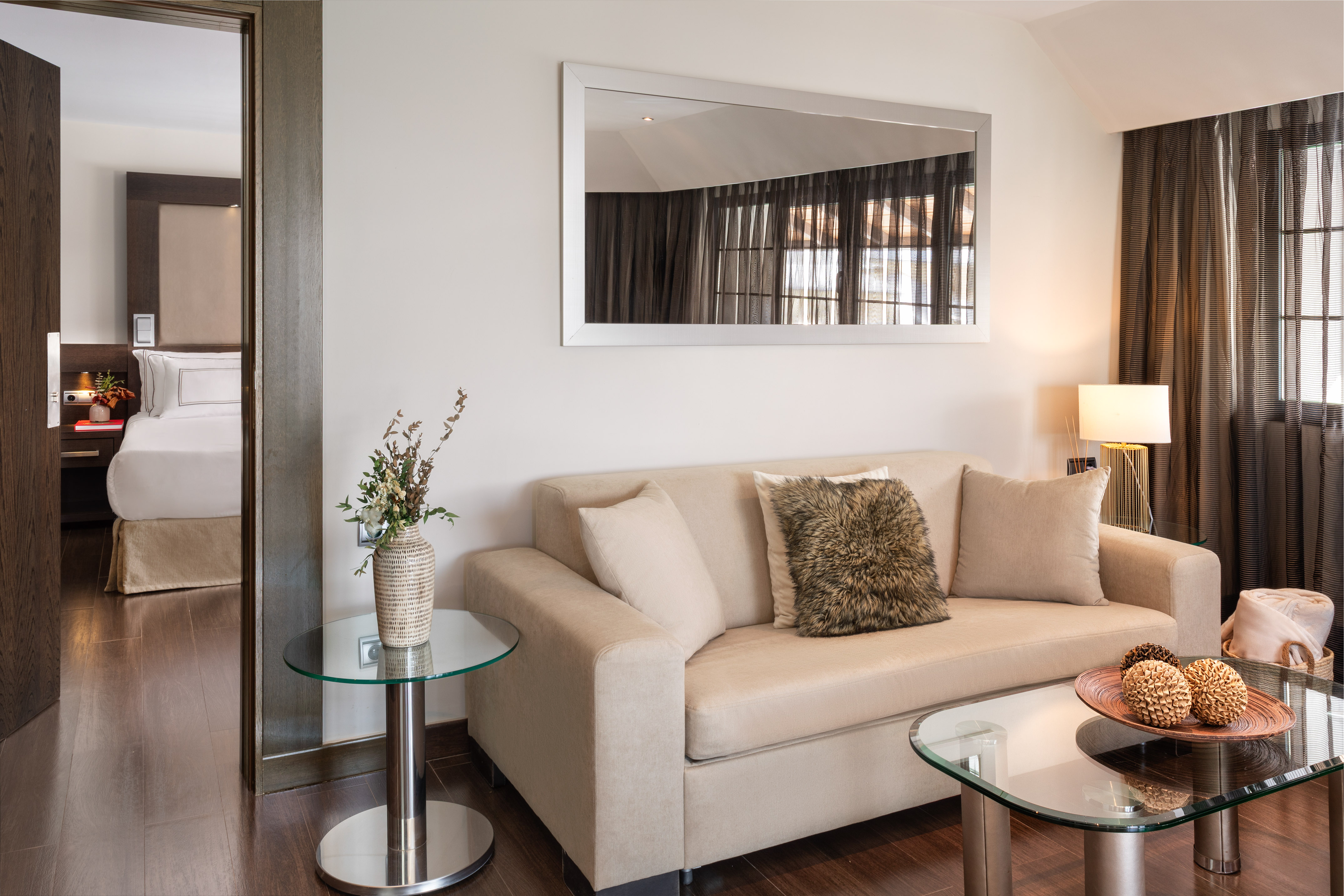 Hotel suite living room with beige sofa, glass tables, mirror, and bedroom entrance.
