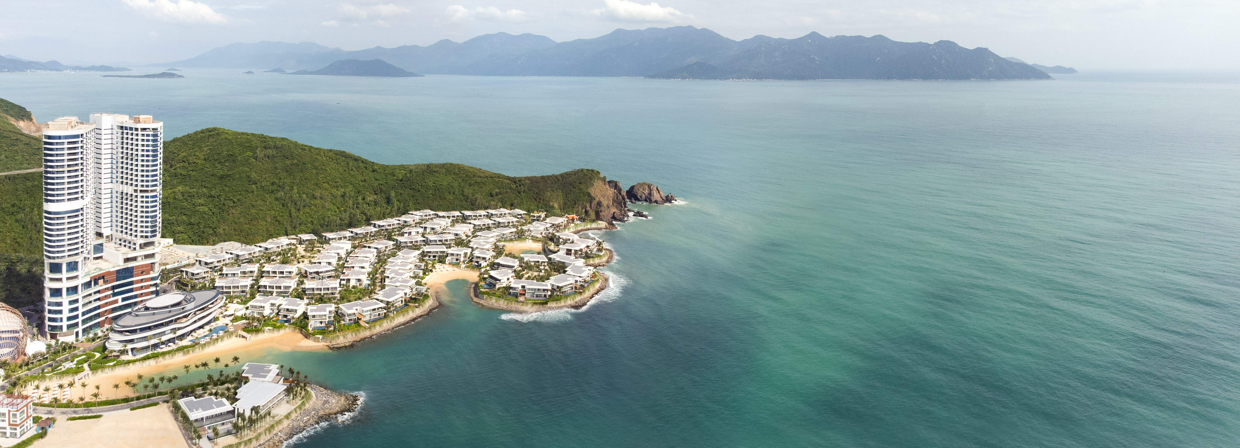 Modern coastal resort and villas on green hills by turquoise ocean.