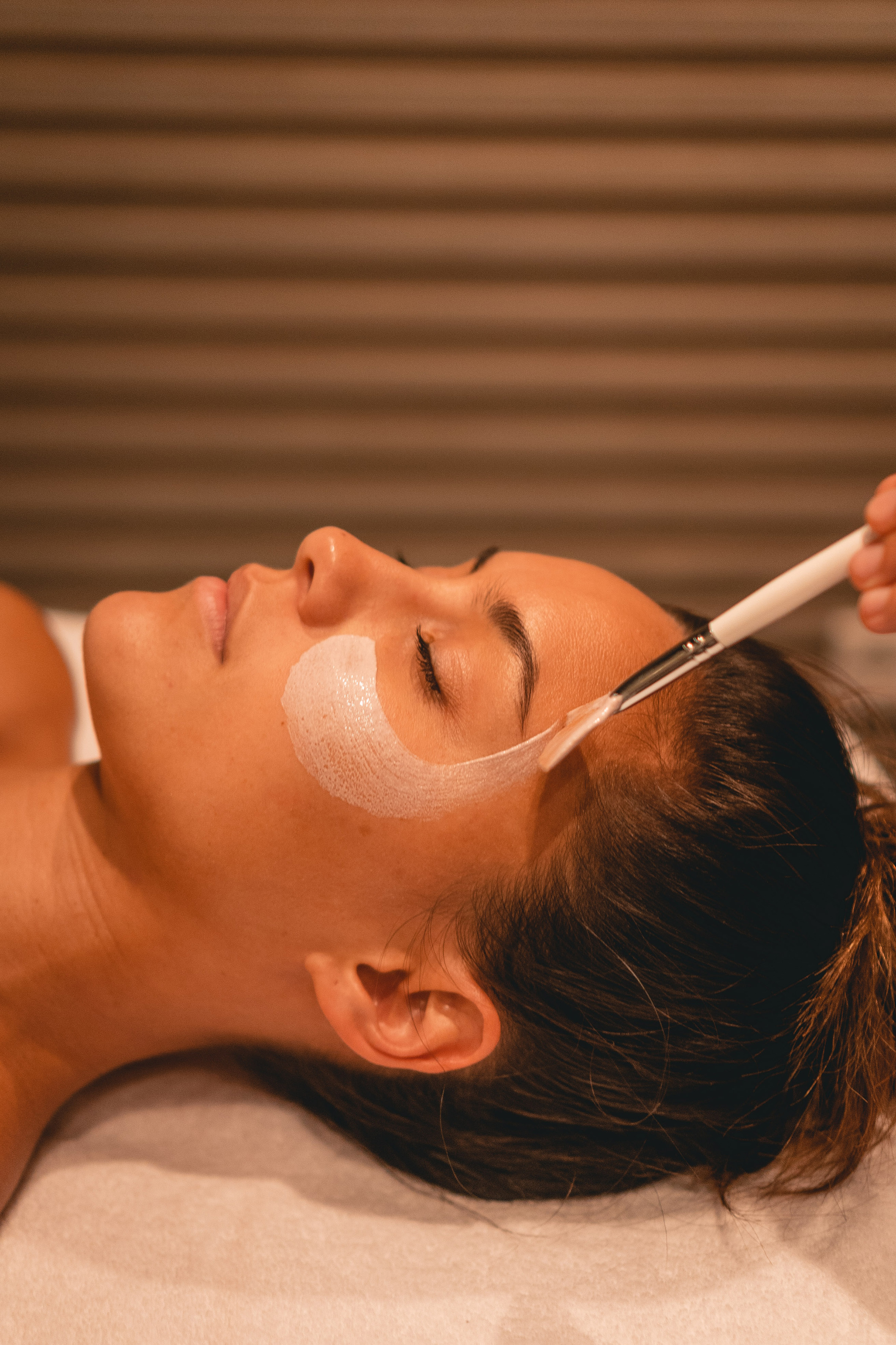 Woman getting white face mask applied with brush during a spa facial.