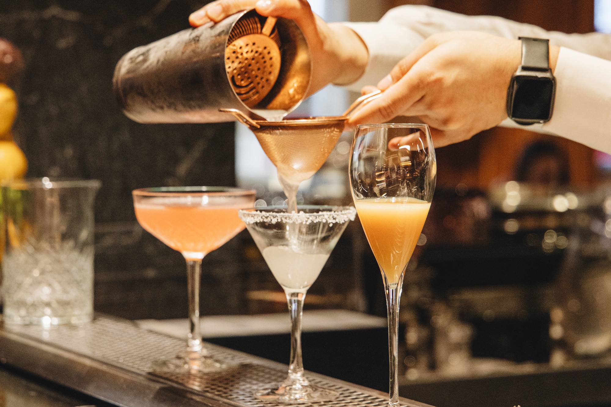 Bartender pouring cocktail from shaker through a strainer into a glass on bar.
