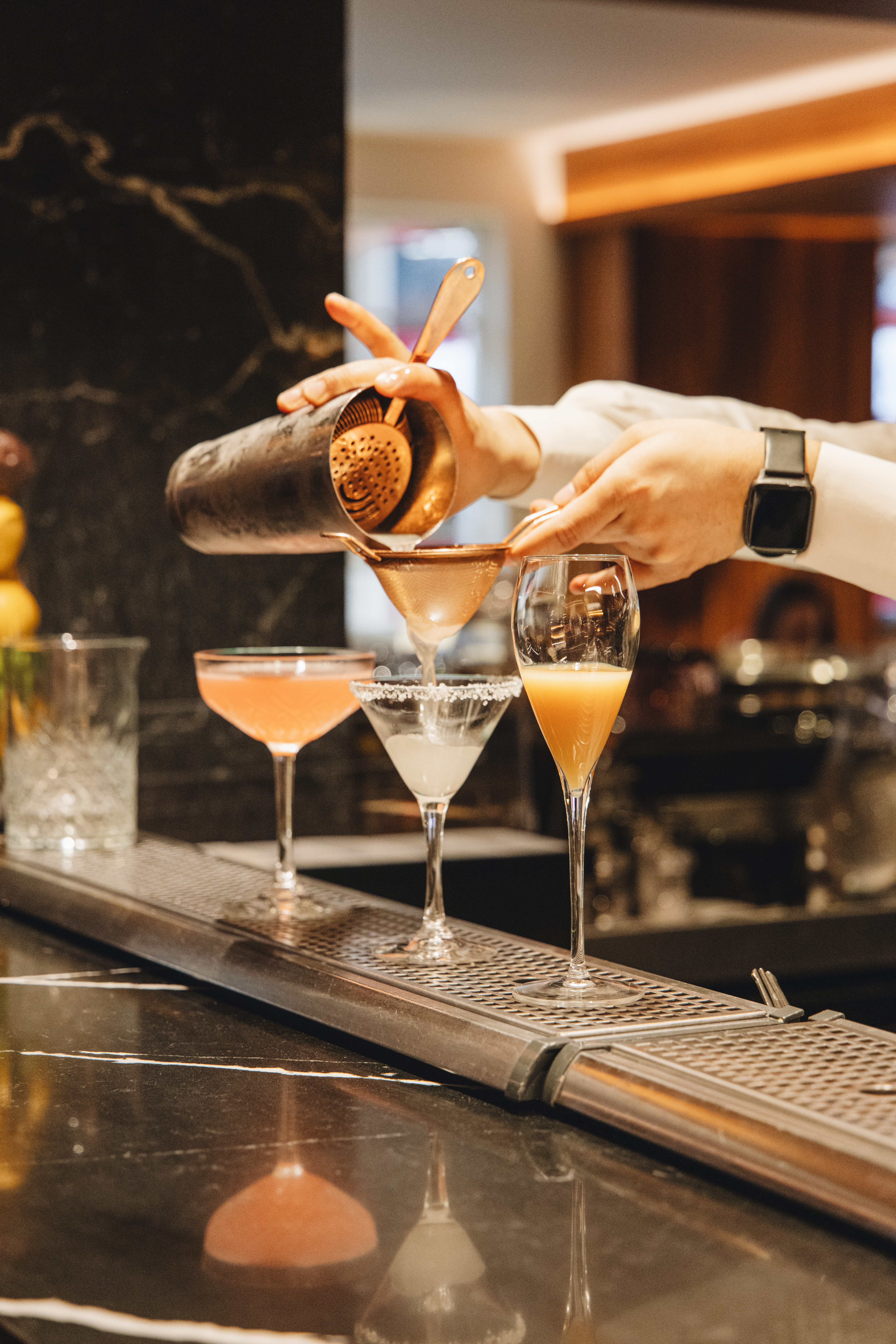 Bartender pouring cocktail from shaker through a strainer into a glass on bar.