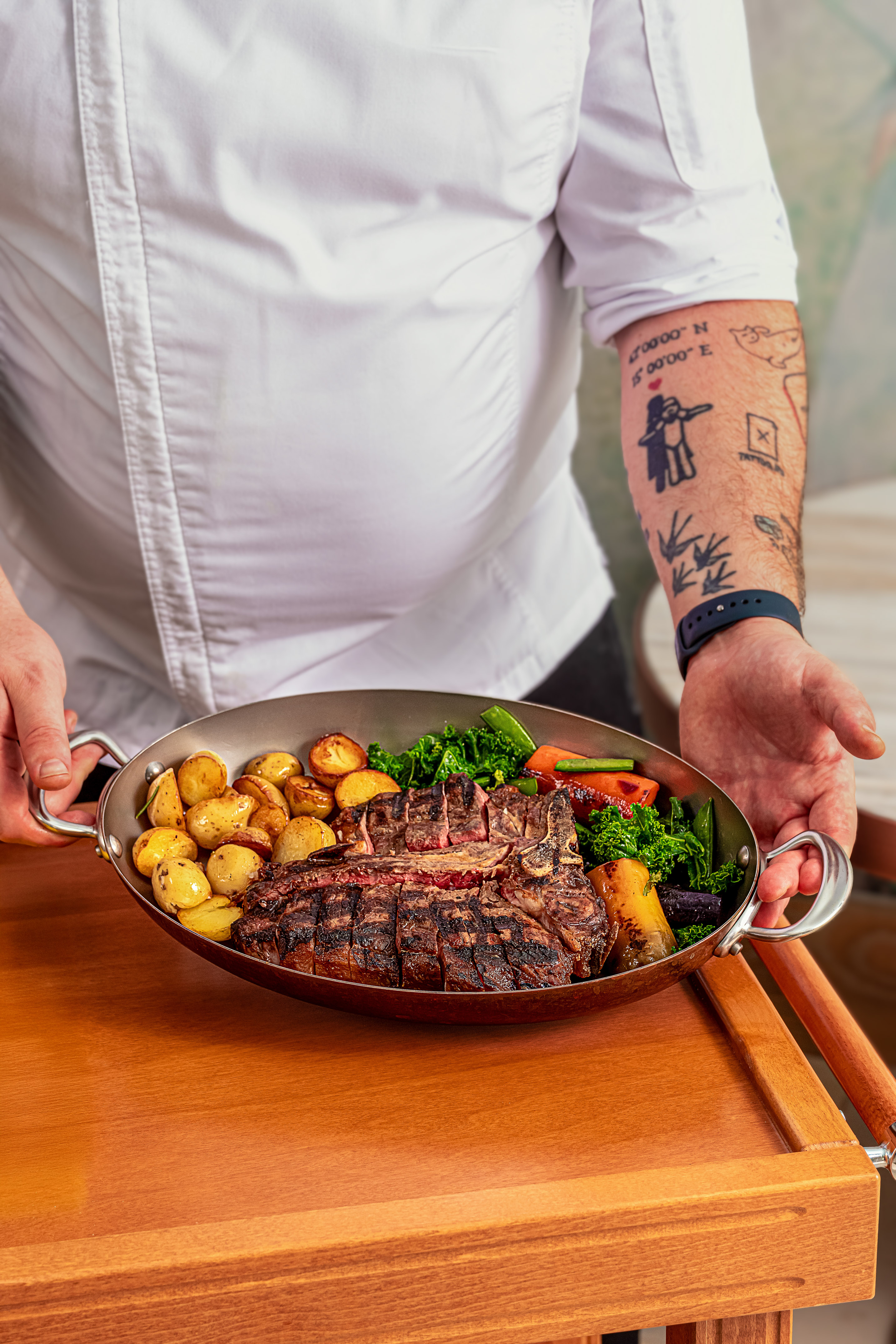 Chef serving grilled bone-in steak, roasted potatoes, and vegetables in skillet.