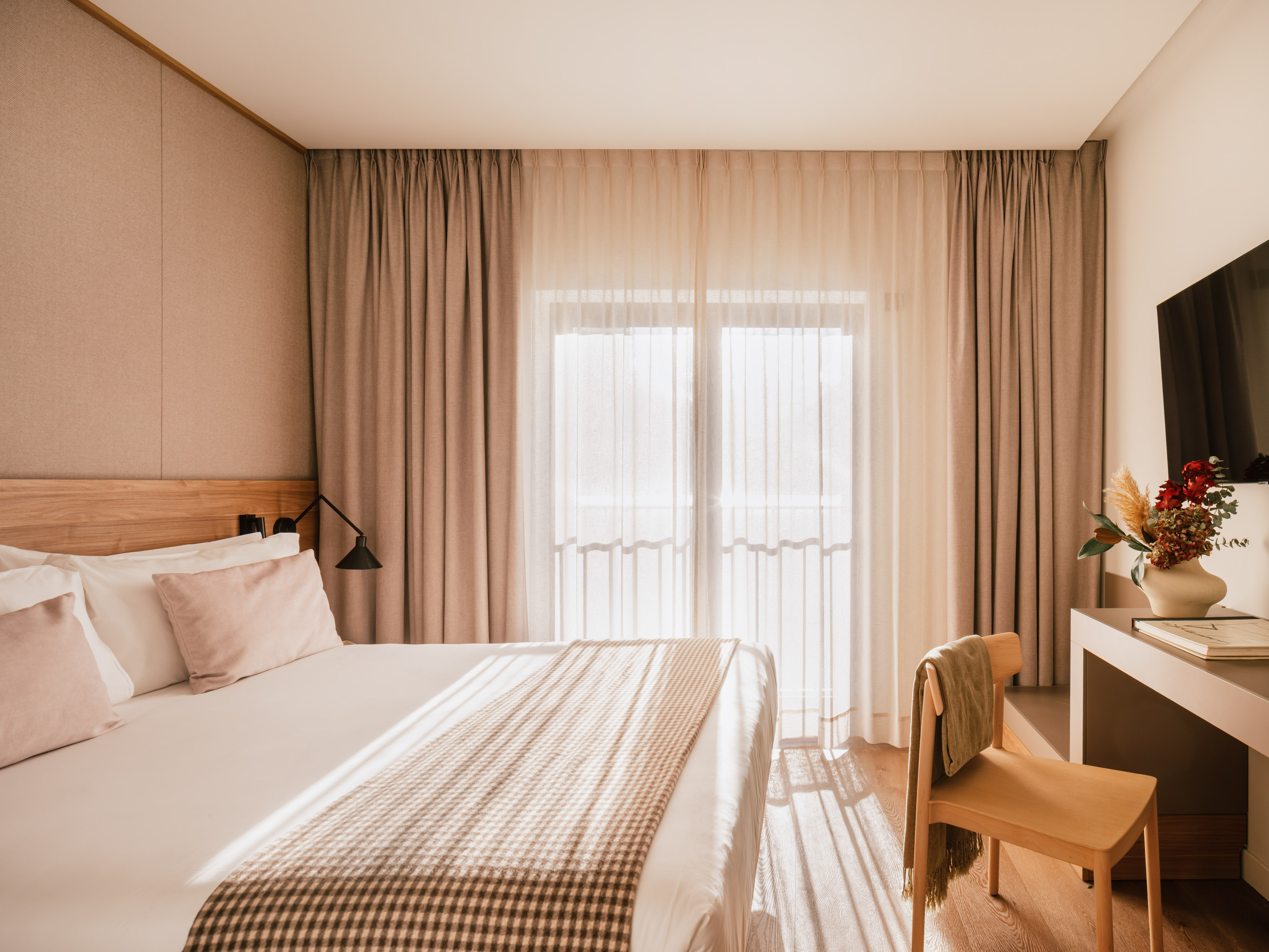 Modern hotel room with bed, bright window, beige curtains, and wooden decor.