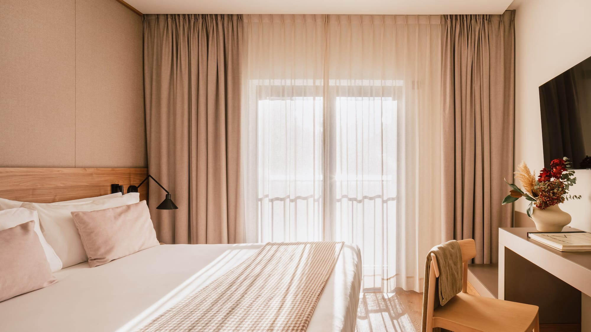 Modern hotel room with bed, bright window, beige curtains, and wooden decor.