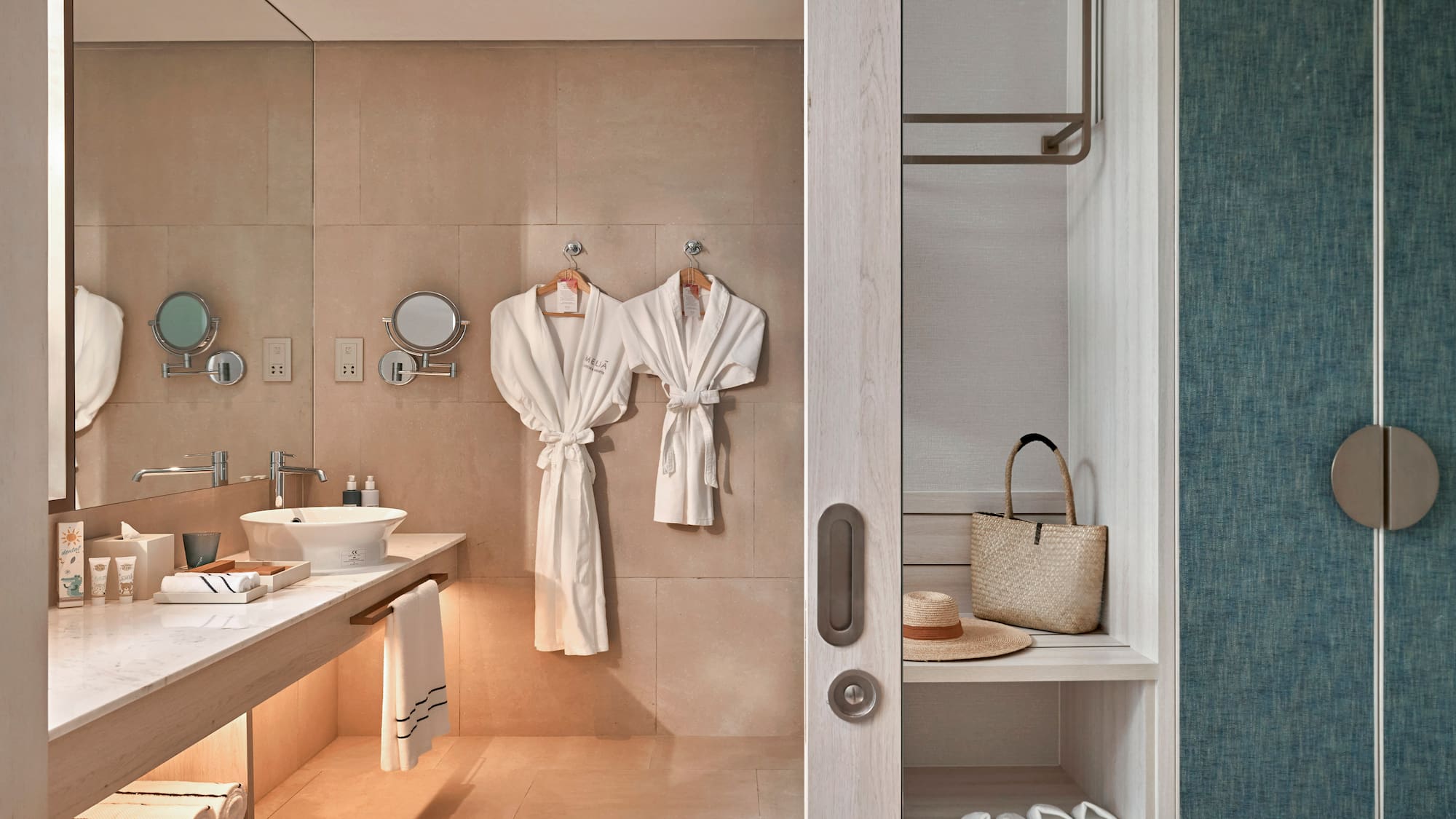 Hotel bathroom with vanity, white bathrobes, and adjacent closet storage.
