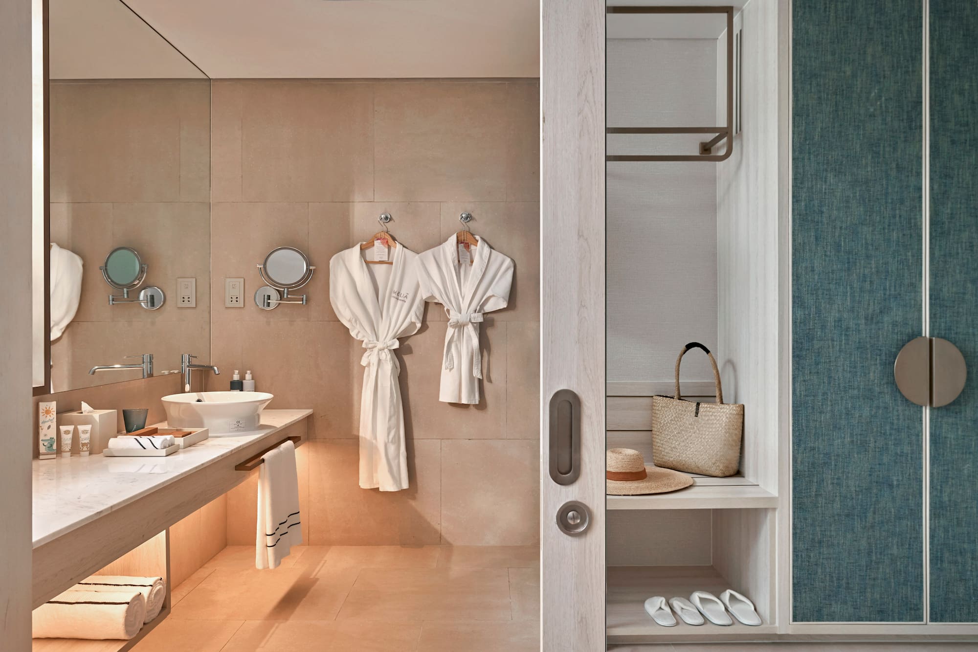 Hotel bathroom with vanity, white bathrobes, and adjacent closet storage.