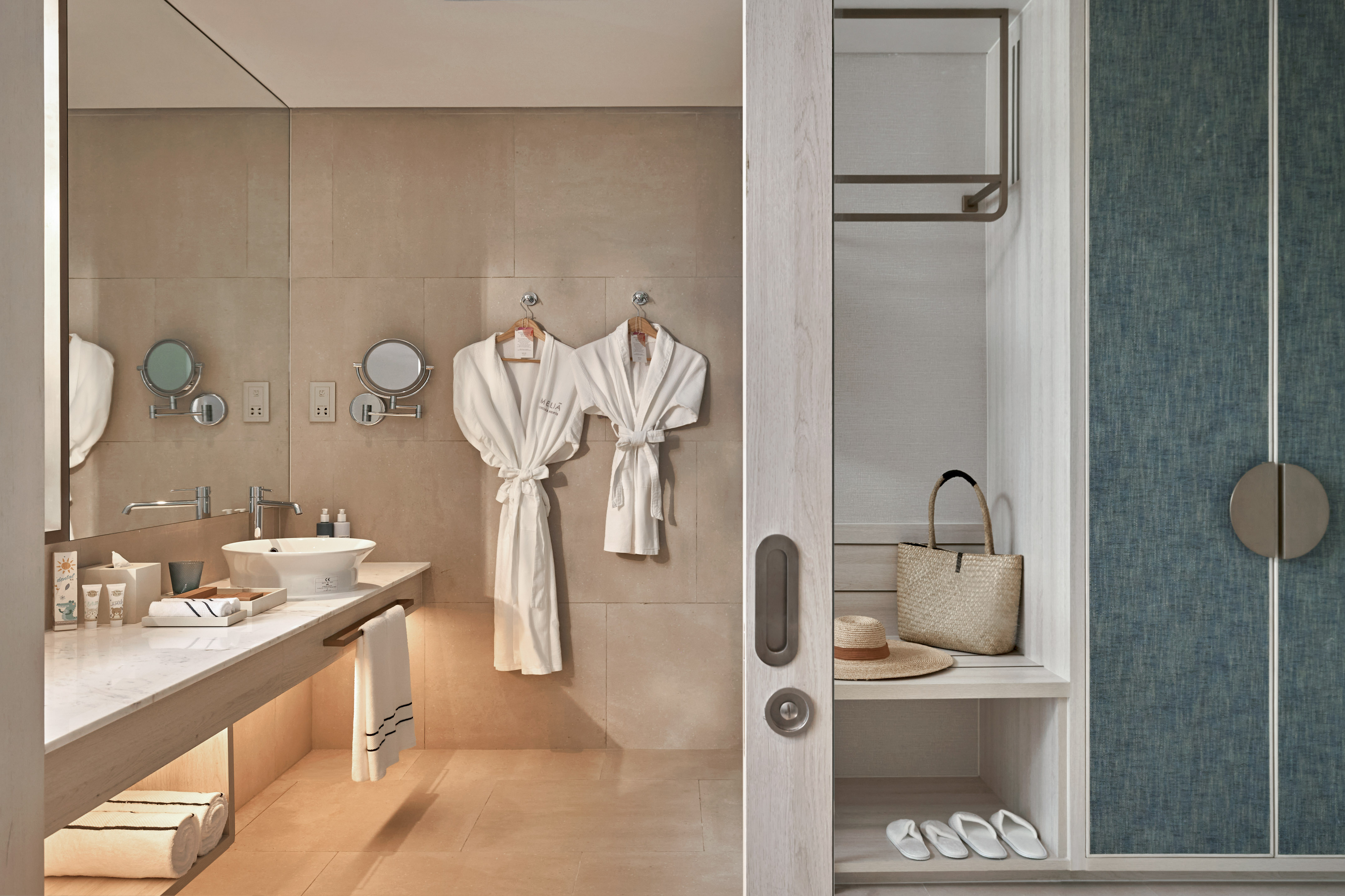 Hotel bathroom with vanity, white bathrobes, and adjacent closet storage.