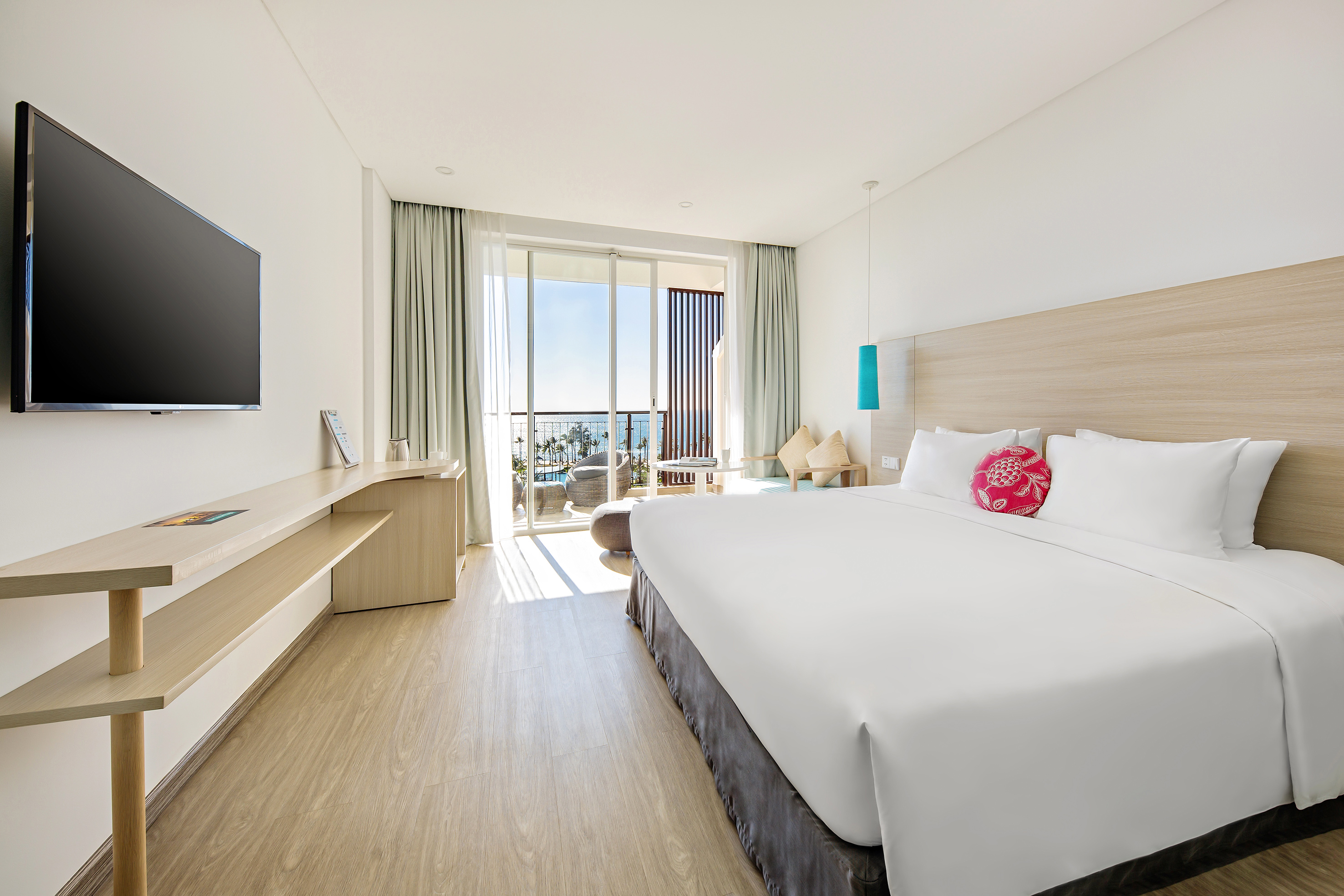 Modern hotel bedroom with large bed, TV, wooden desk, and balcony ocean view.