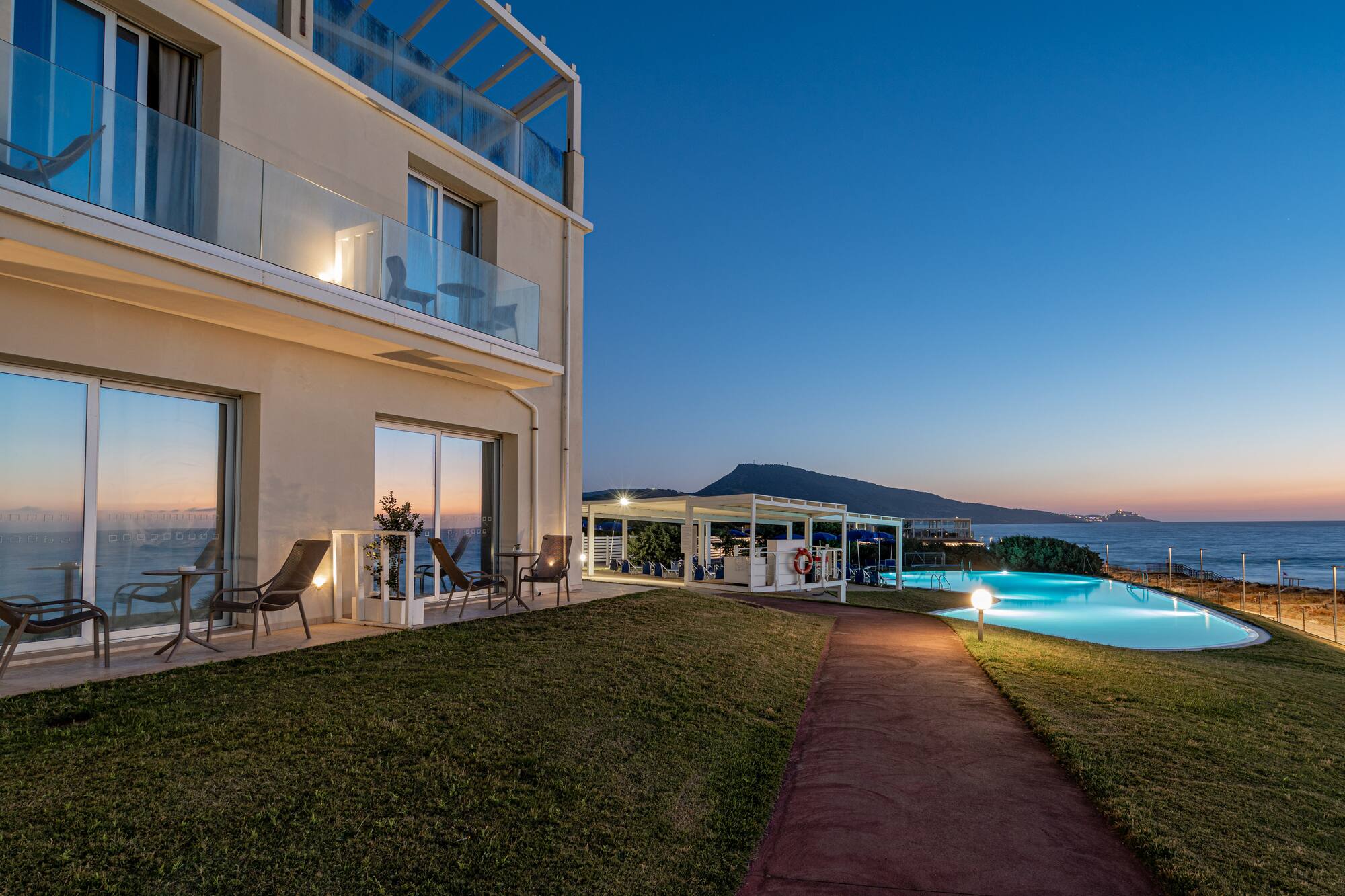 Beachfront hotel with illuminated swimming pool and ocean view at dusk.