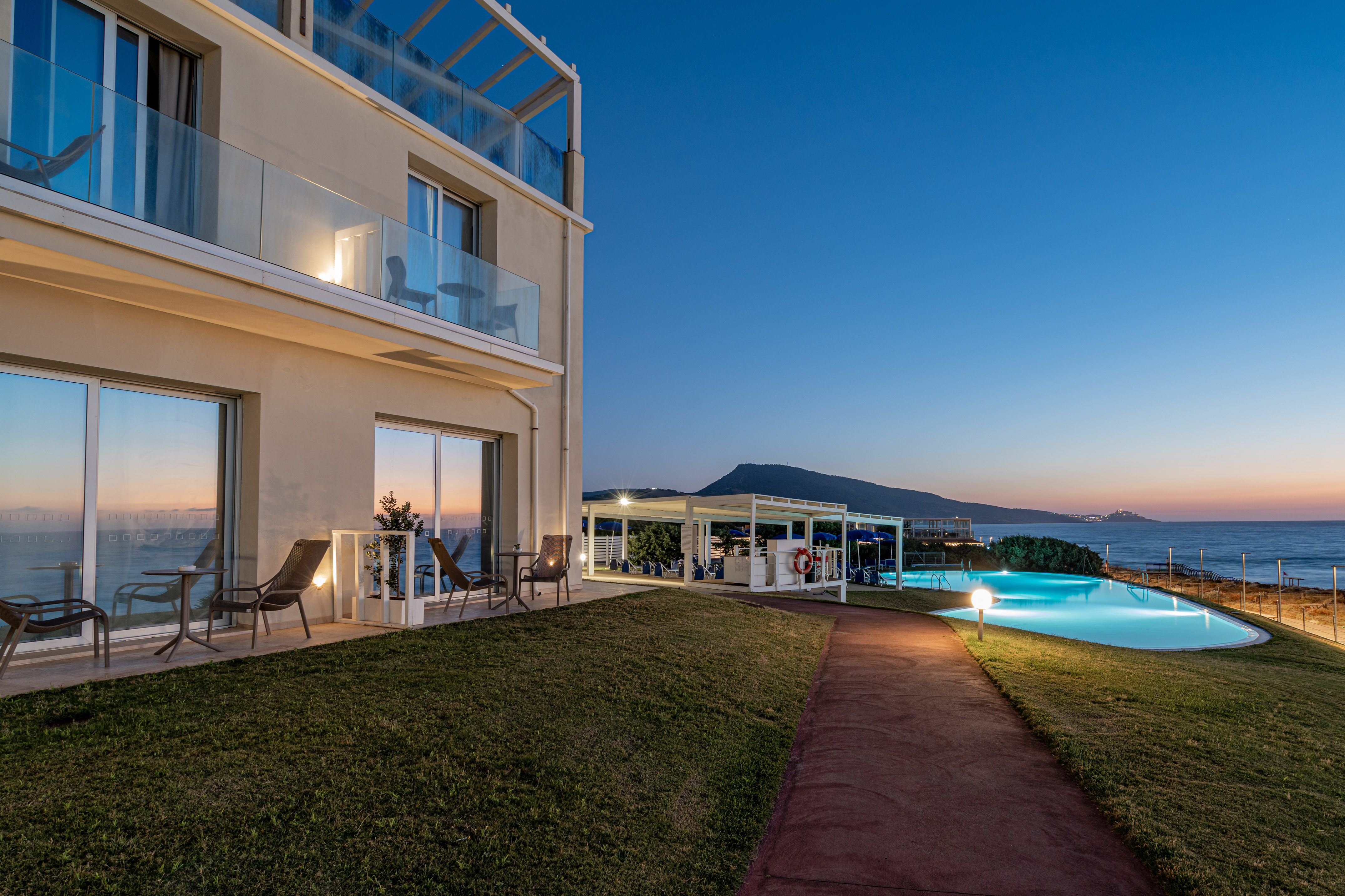 Beachfront hotel with illuminated swimming pool and ocean view at dusk.