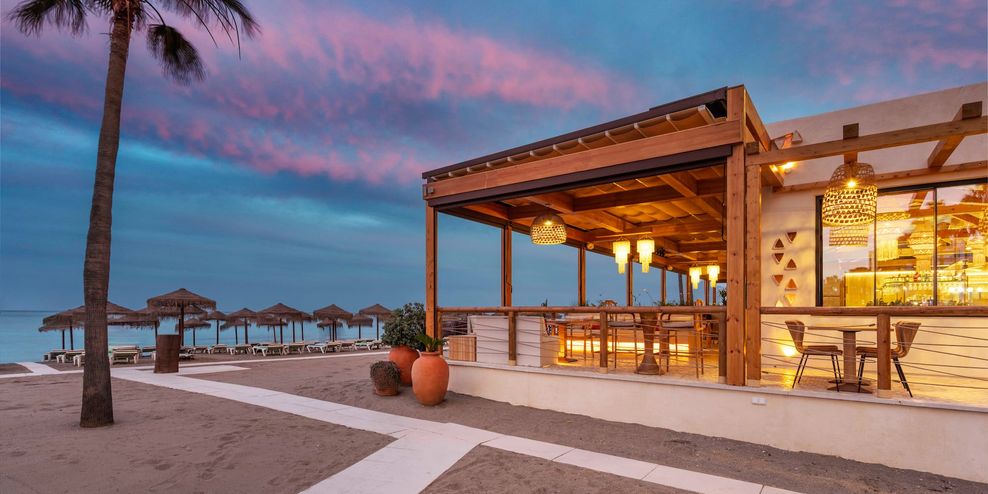 Illuminated beachfront restaurant with outdoor seating, sandy beach, and twilight sky.