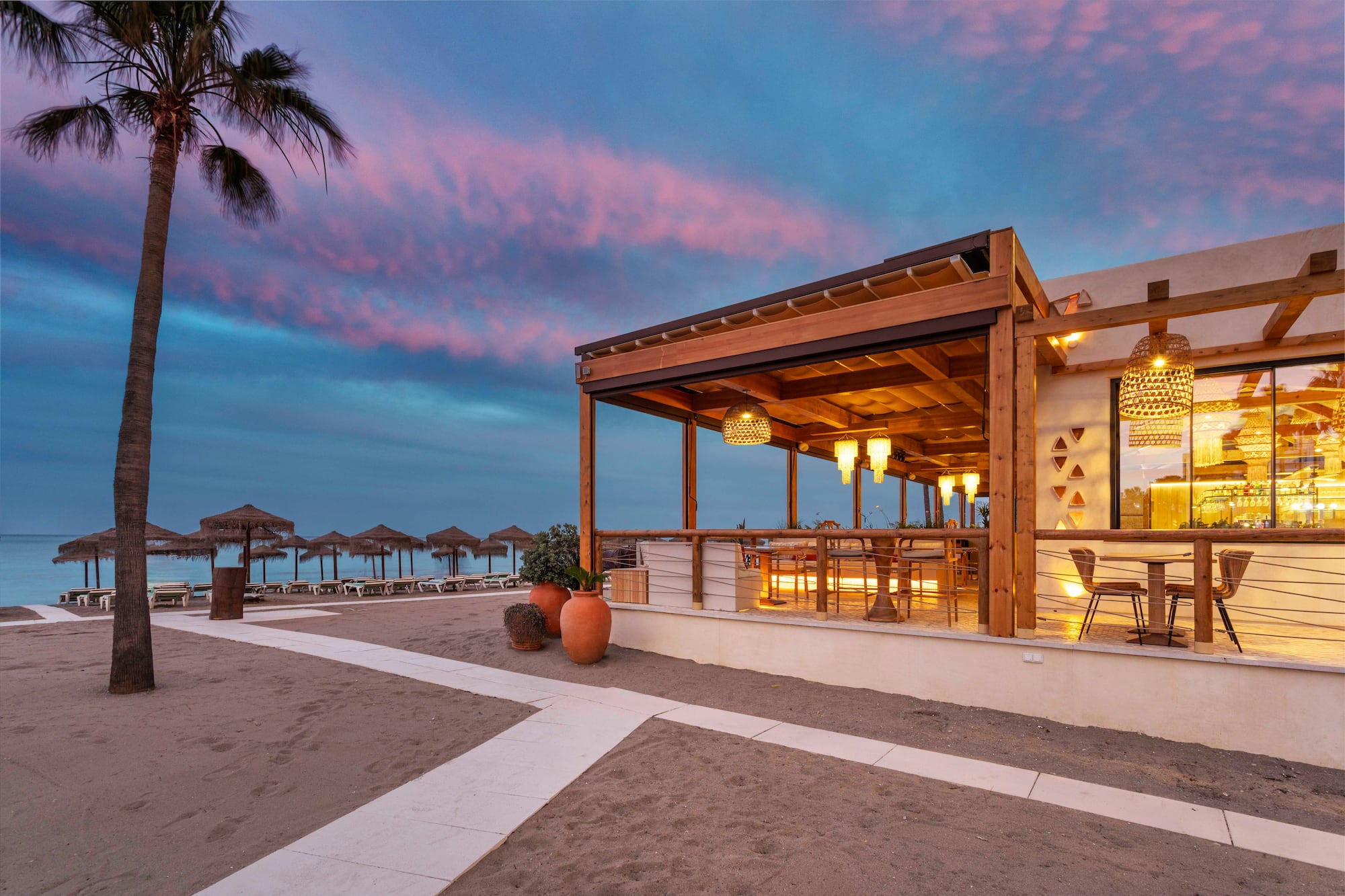 Illuminated beachfront restaurant with outdoor seating, sandy beach, and twilight sky.
