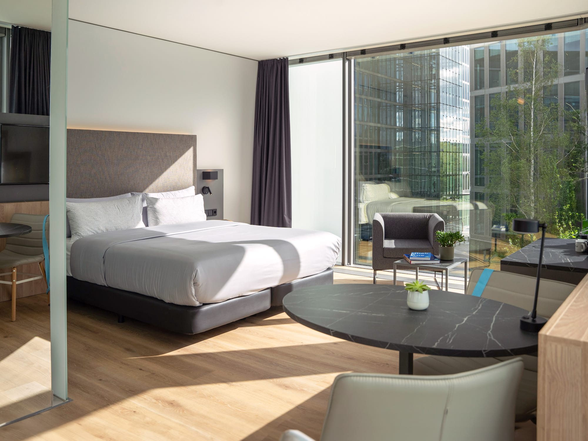 Modern hotel room with bed, large window city view, wooden floor, and dining area.