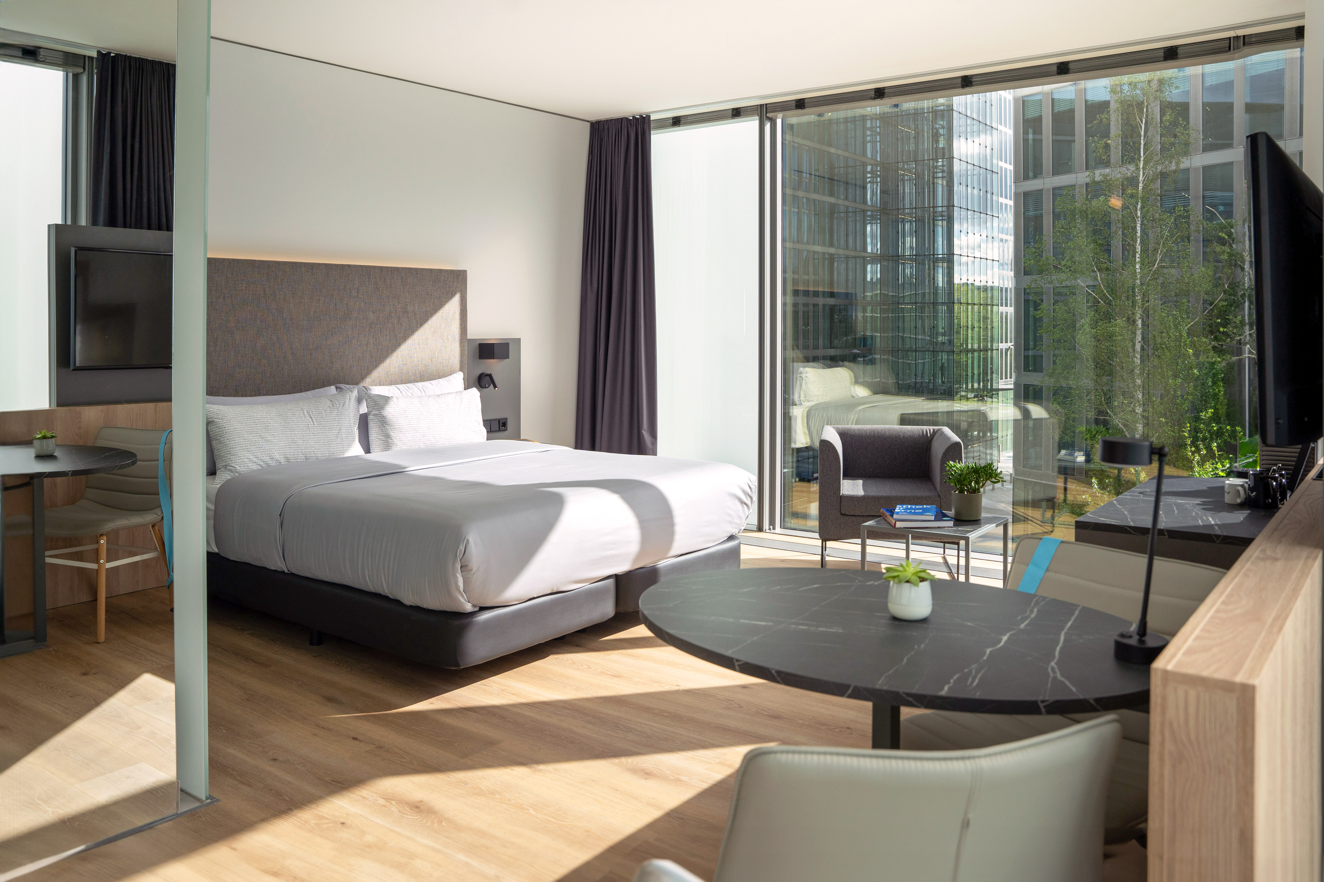 Modern hotel room with bed, large window city view, wooden floor, and dining area.