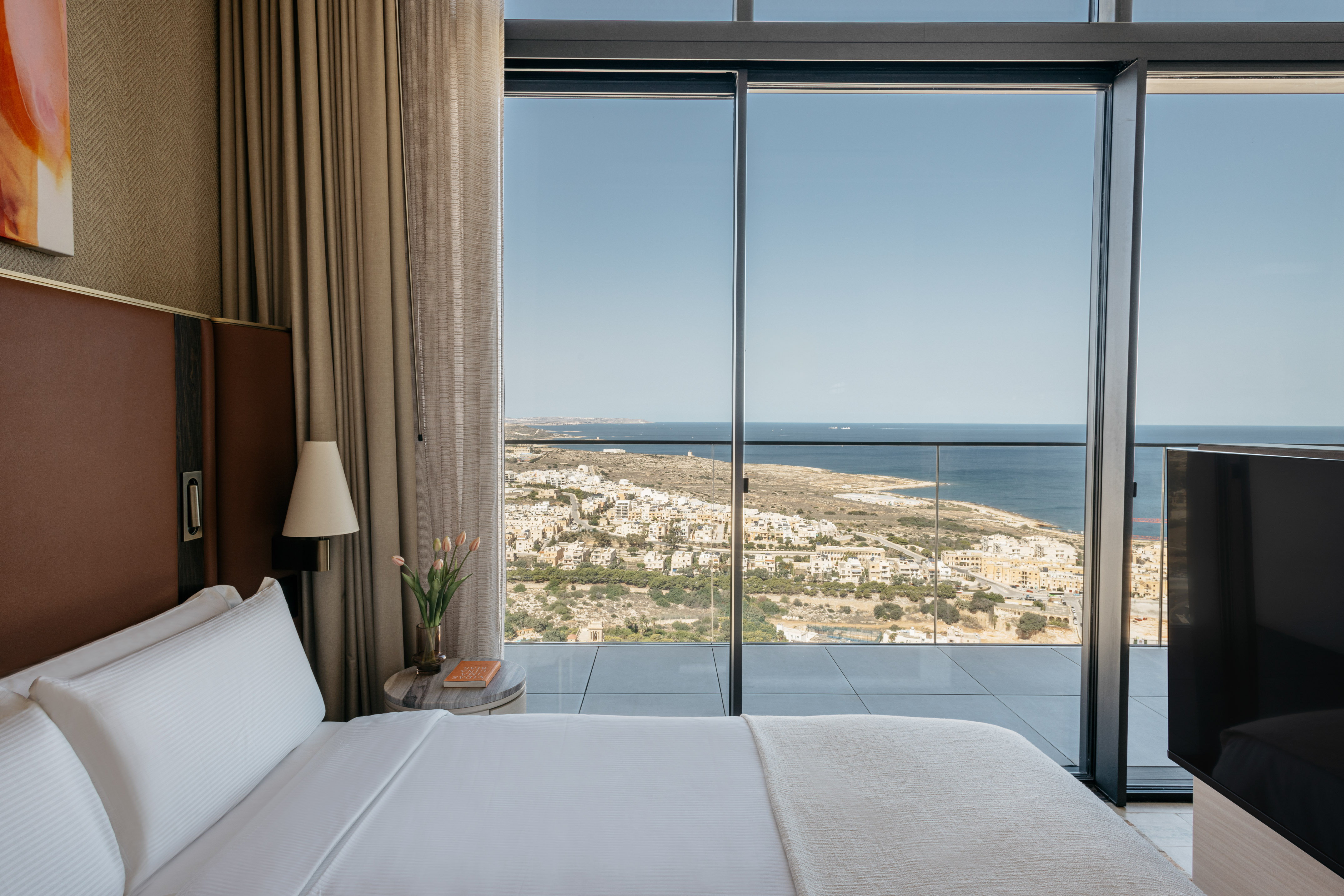 Modern bedroom with white bed, panoramic ocean view, and coastal town.