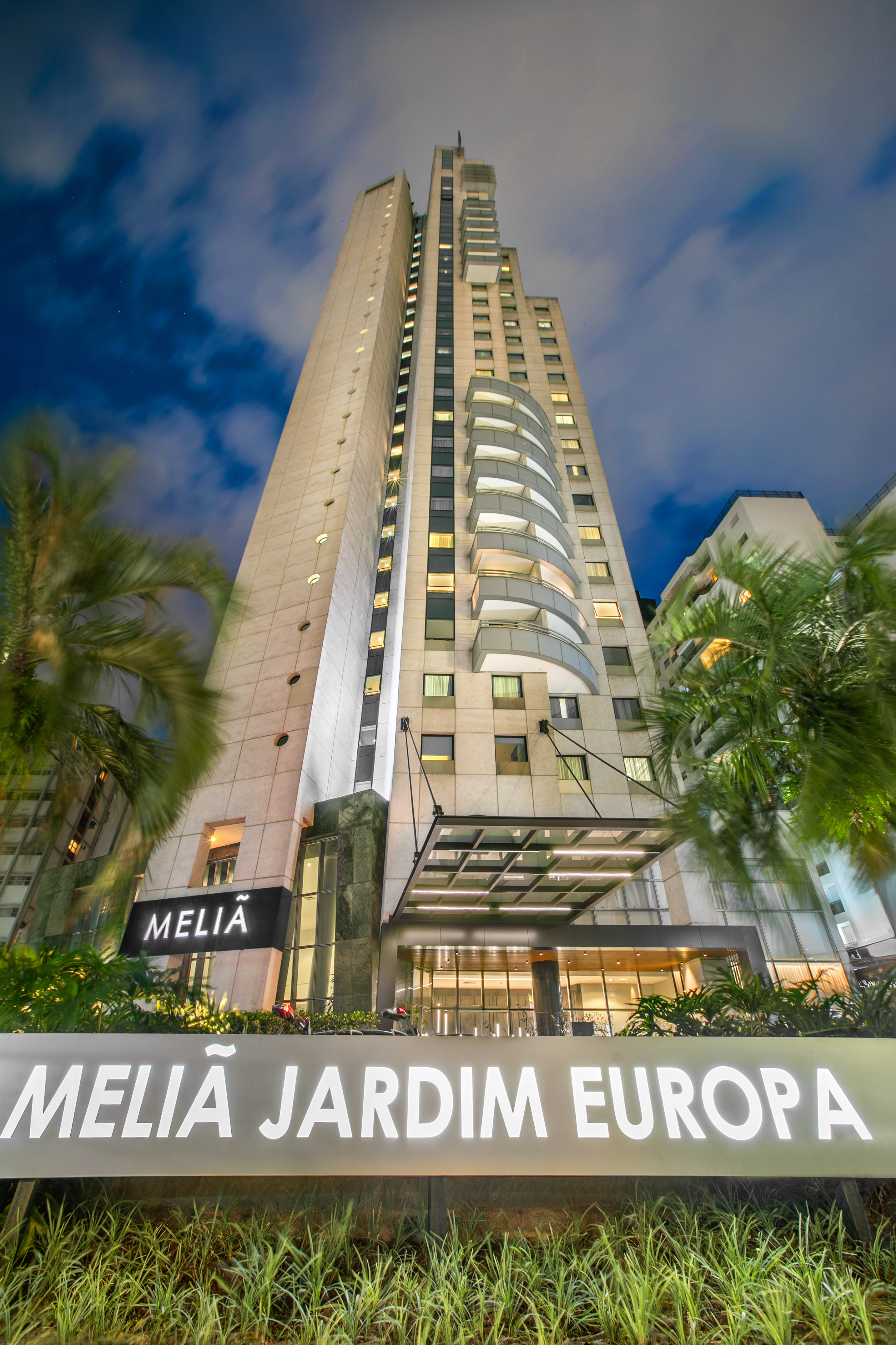 Melia Jardim Europa hotel skyscraper at night, illuminated sign, palm trees.
