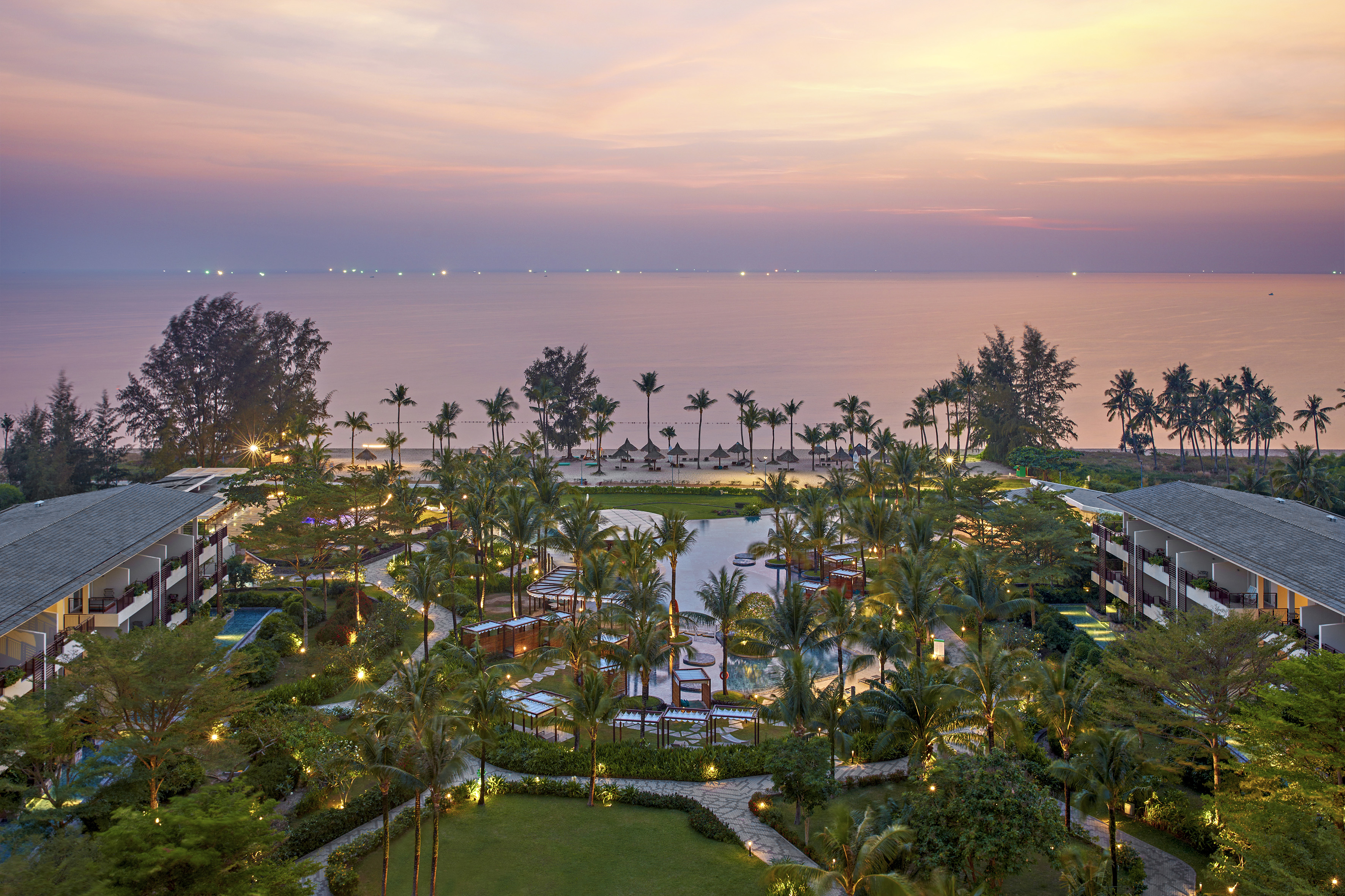 Aerial view of tropical beach resort, pool, palm trees, ocean at sunset.