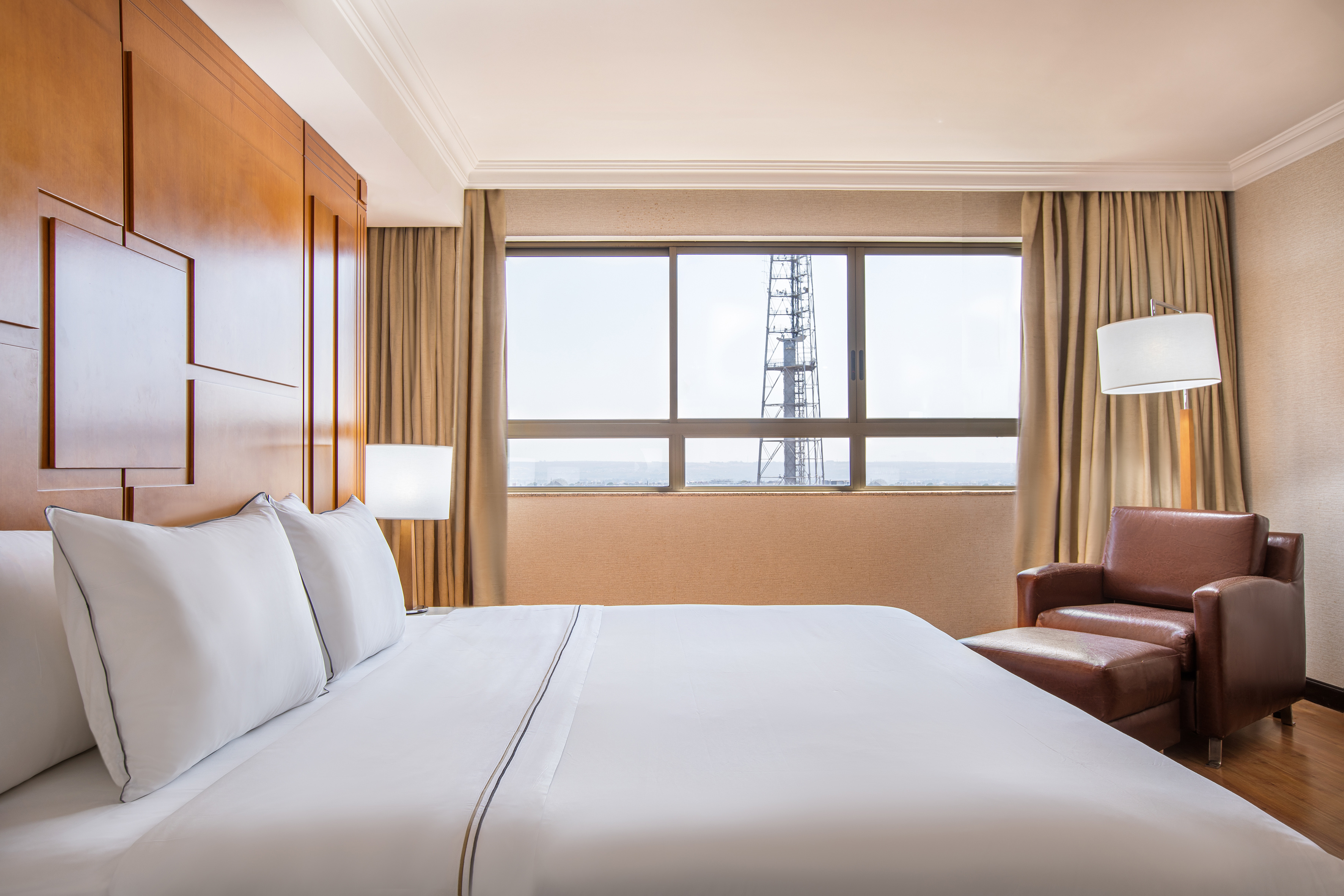 Hotel bedroom with white bed, wood paneling, leather chair, and industrial tower view.