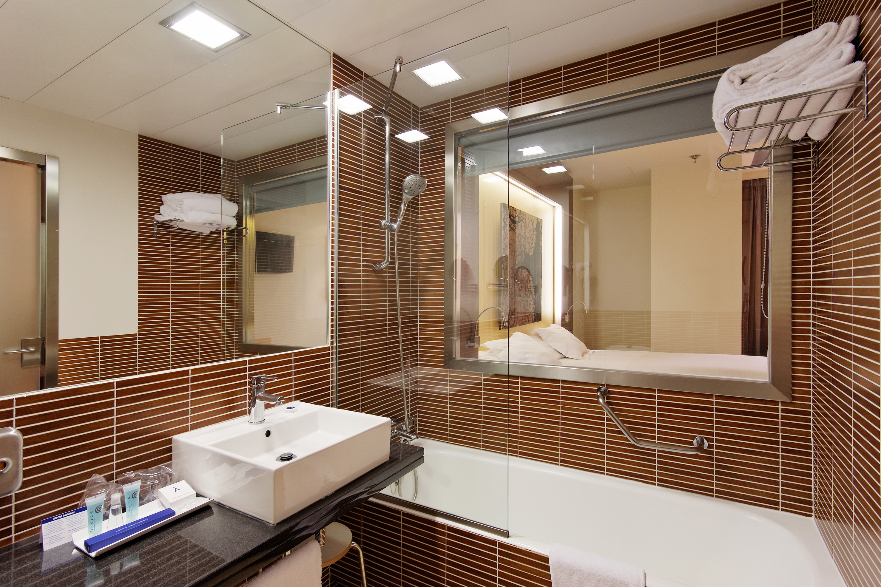 Modern hotel bathroom with brown tiles, white sink, tub, and window to bedroom.