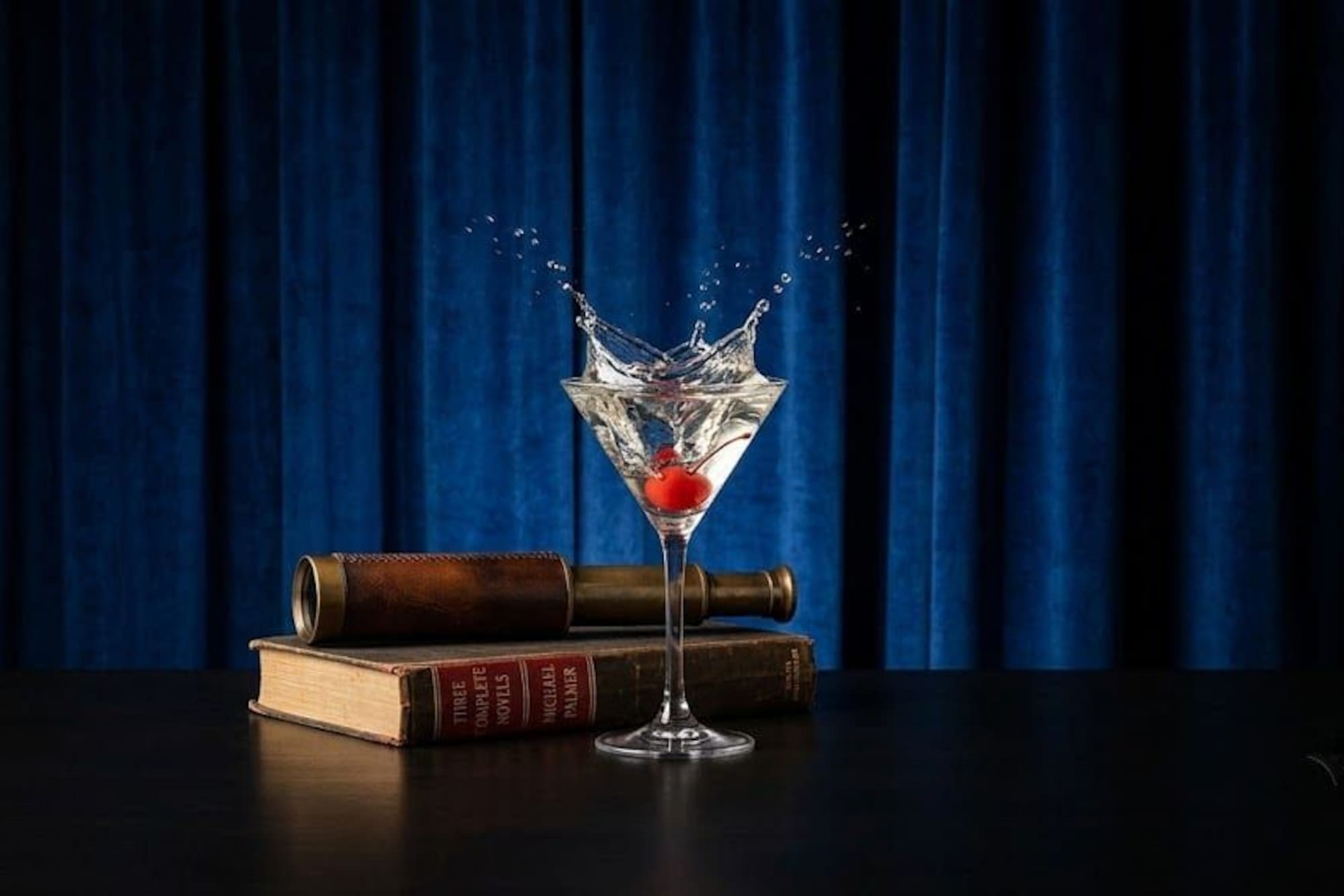 Elegant martini splash with cherry, vintage book, telescope, blue curtains.