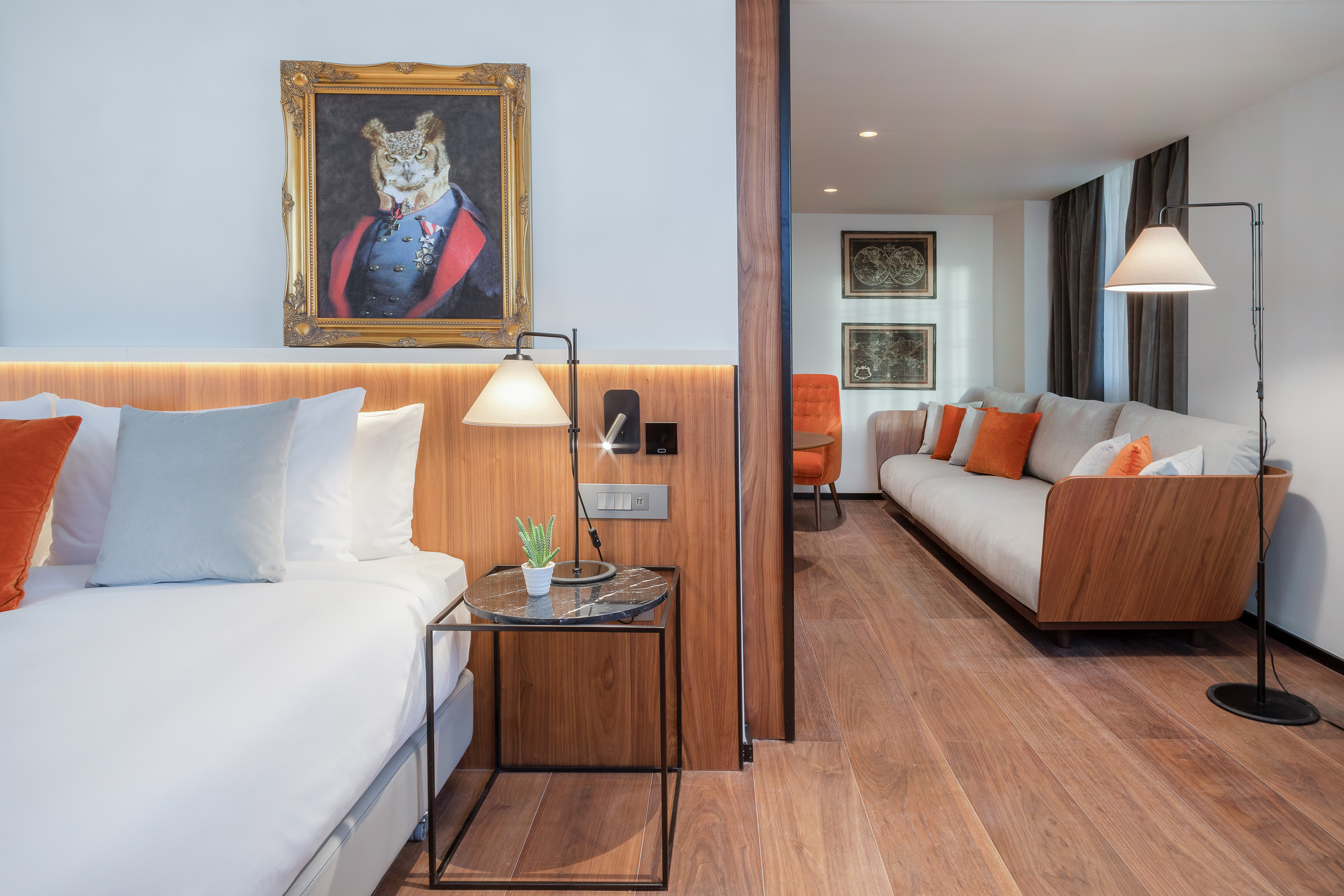 Modern hotel suite with bed, sofa, wood floor, and distinct owl painting.