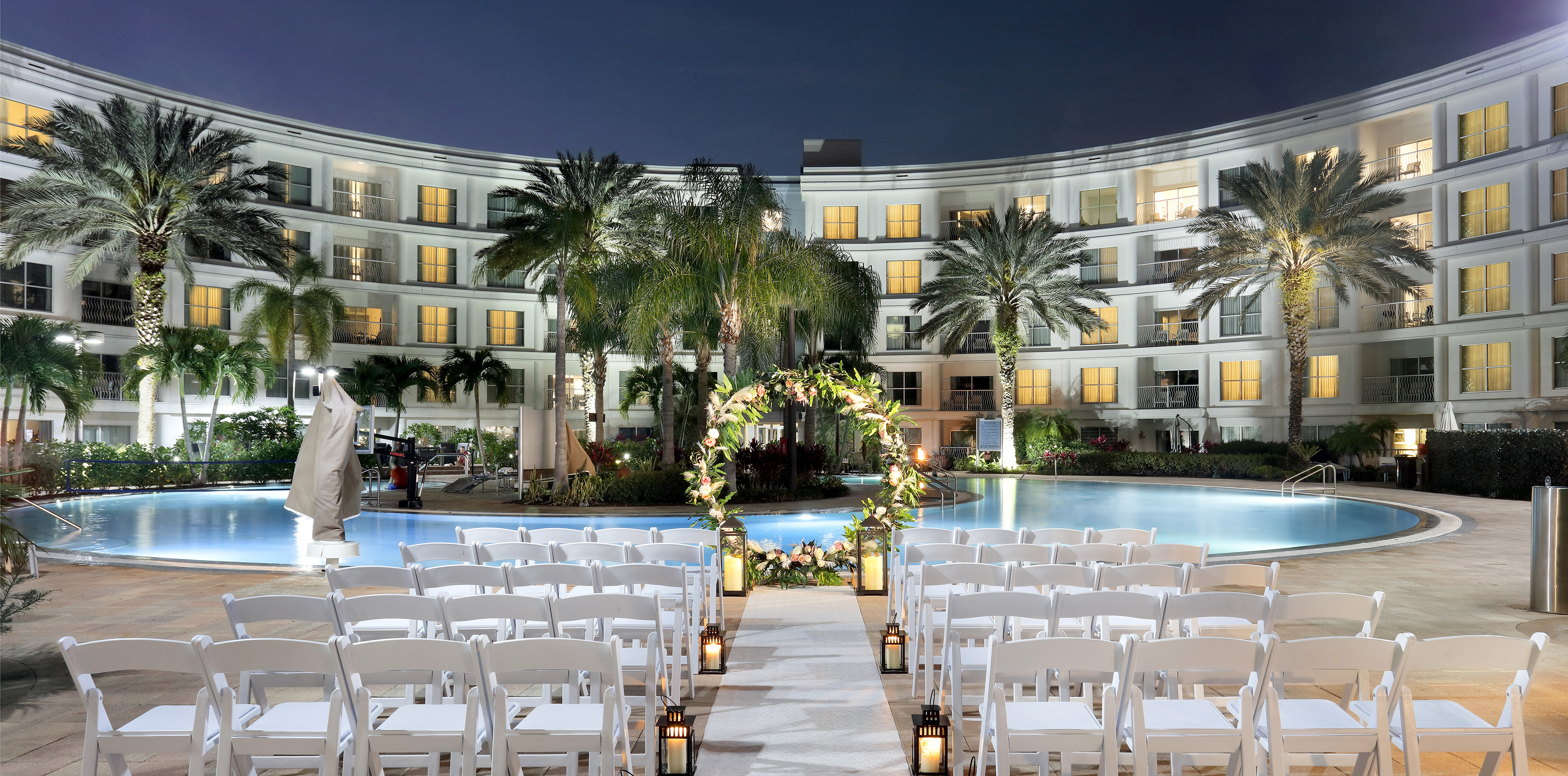 Tropical resort wedding ceremony at night by pool, white chairs, floral arch. - Pool Side (outdoors) - Meliá Orlando Celebration, Orlando