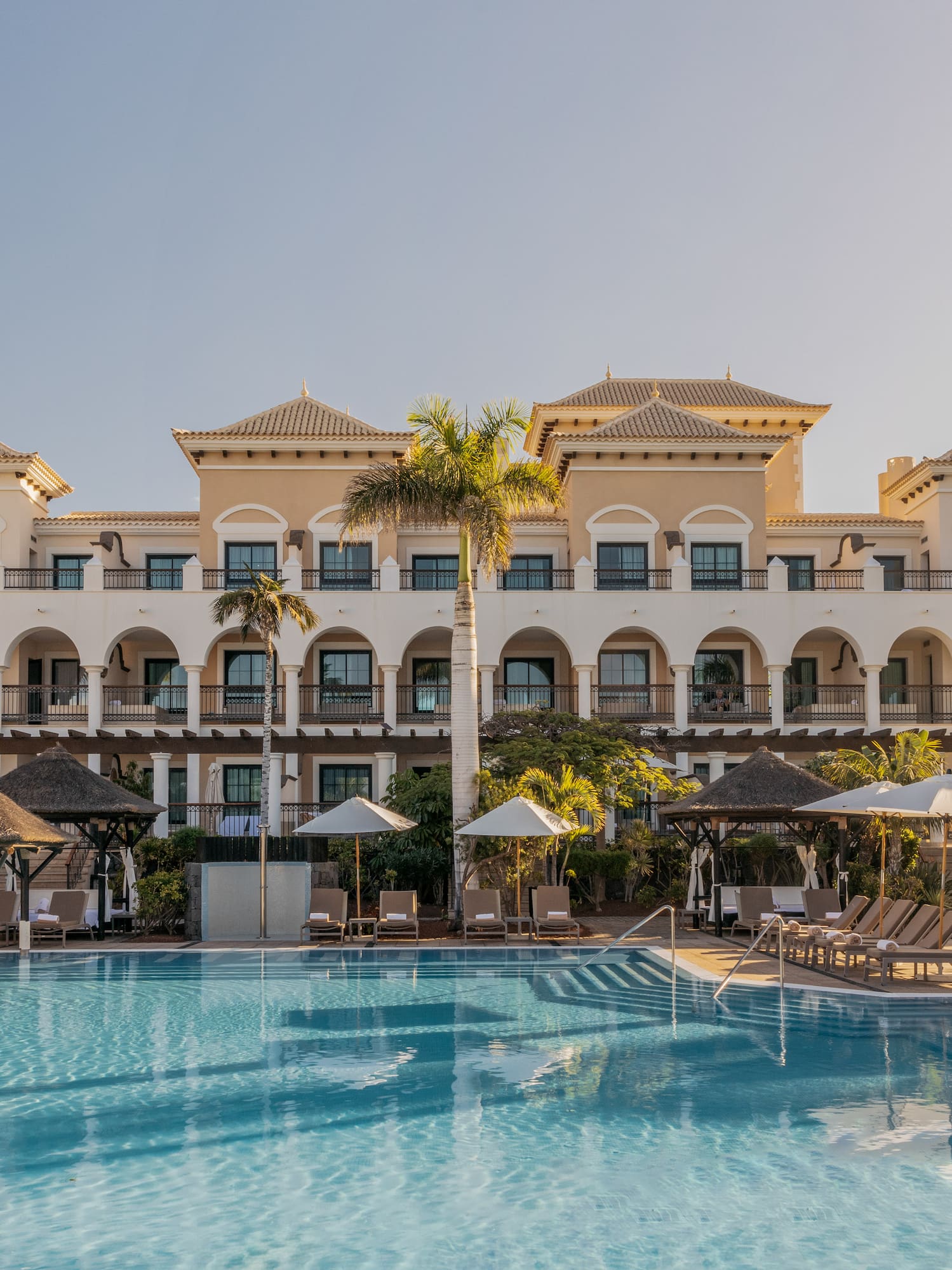 Mediterranean resort hotel with large pool, lounge chairs, and palm trees.