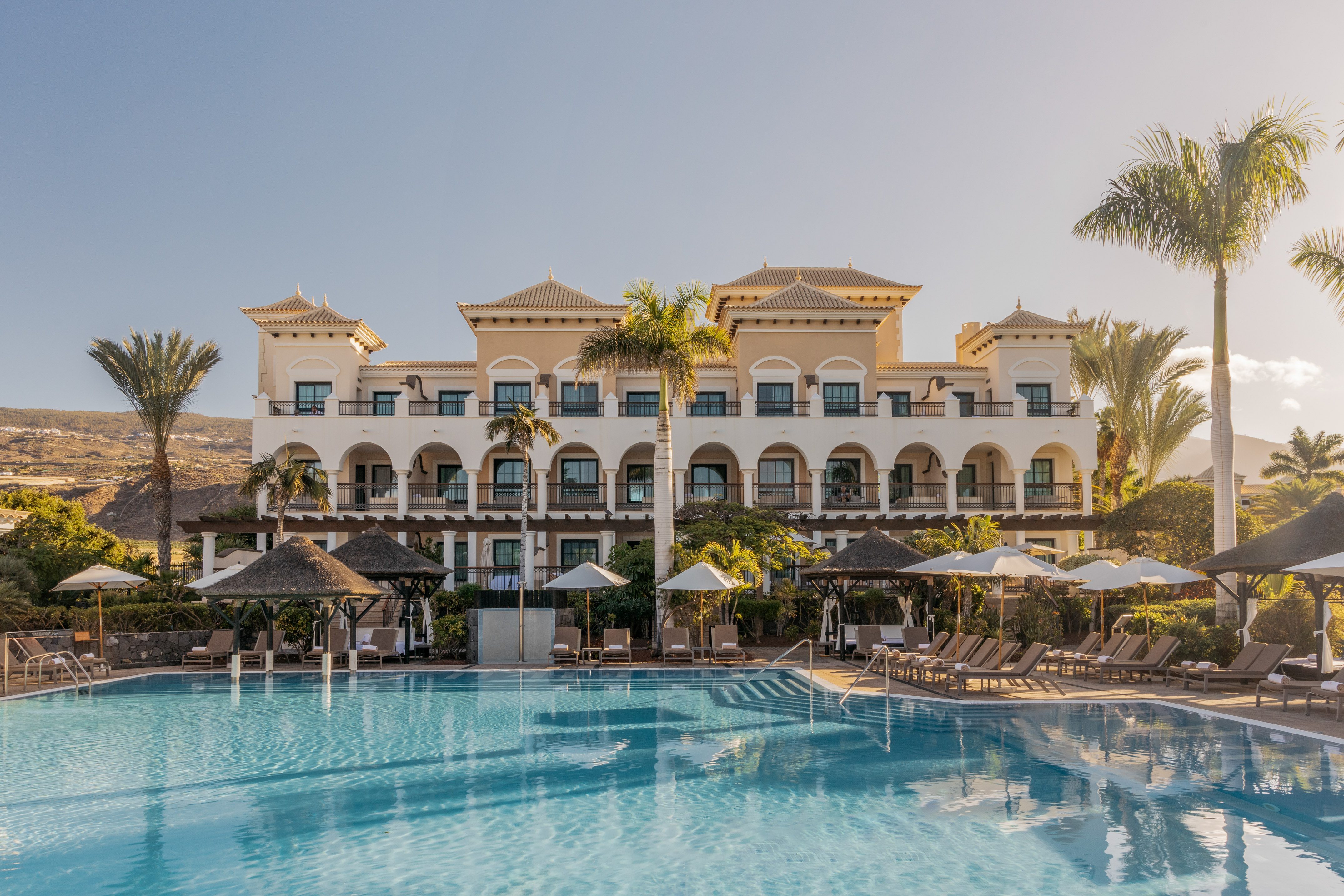 Mediterranean resort hotel with large pool, lounge chairs, and palm trees.