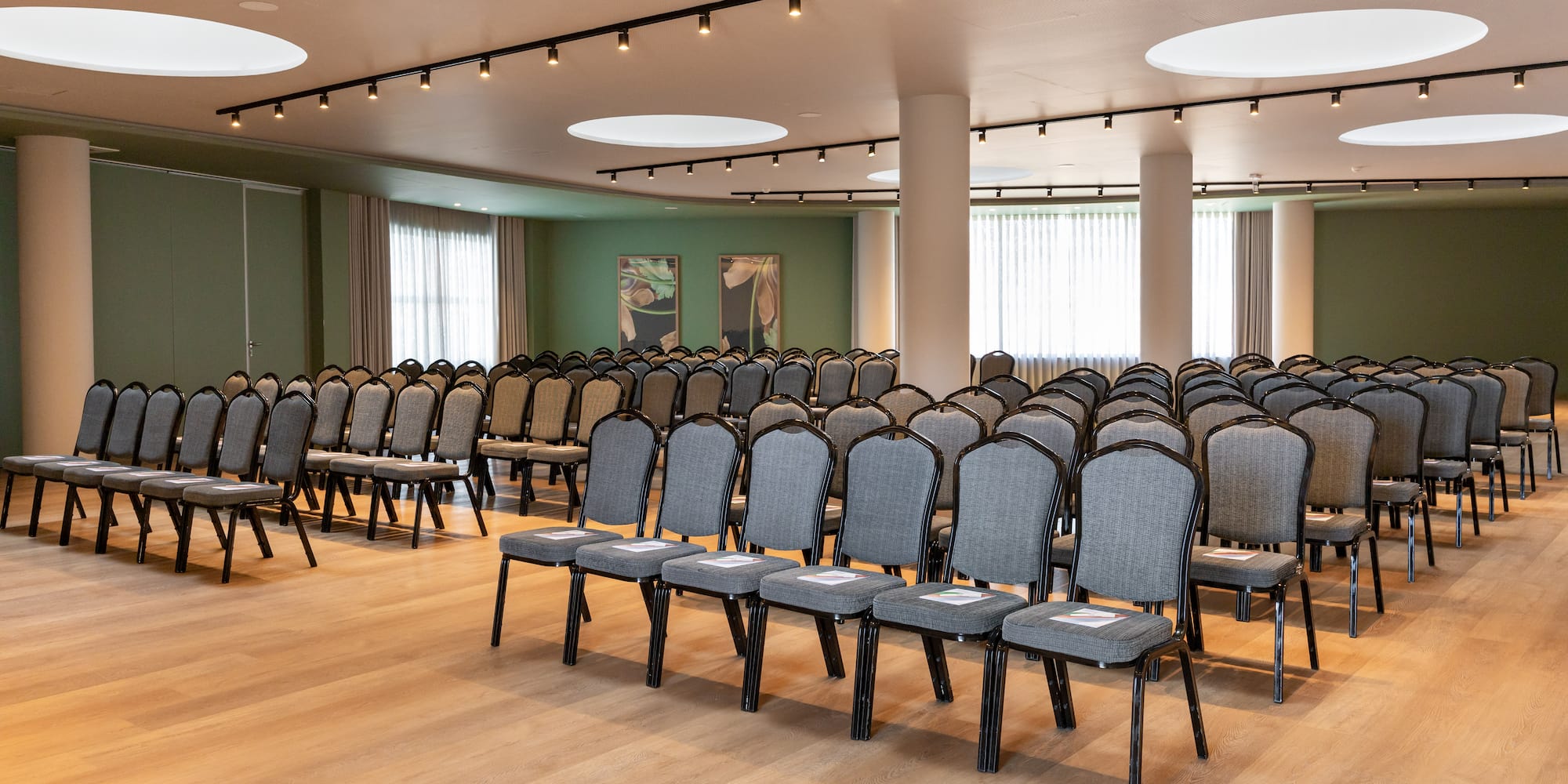 Spacious empty conference room with rows of grey chairs, hardwood floor, and green walls.