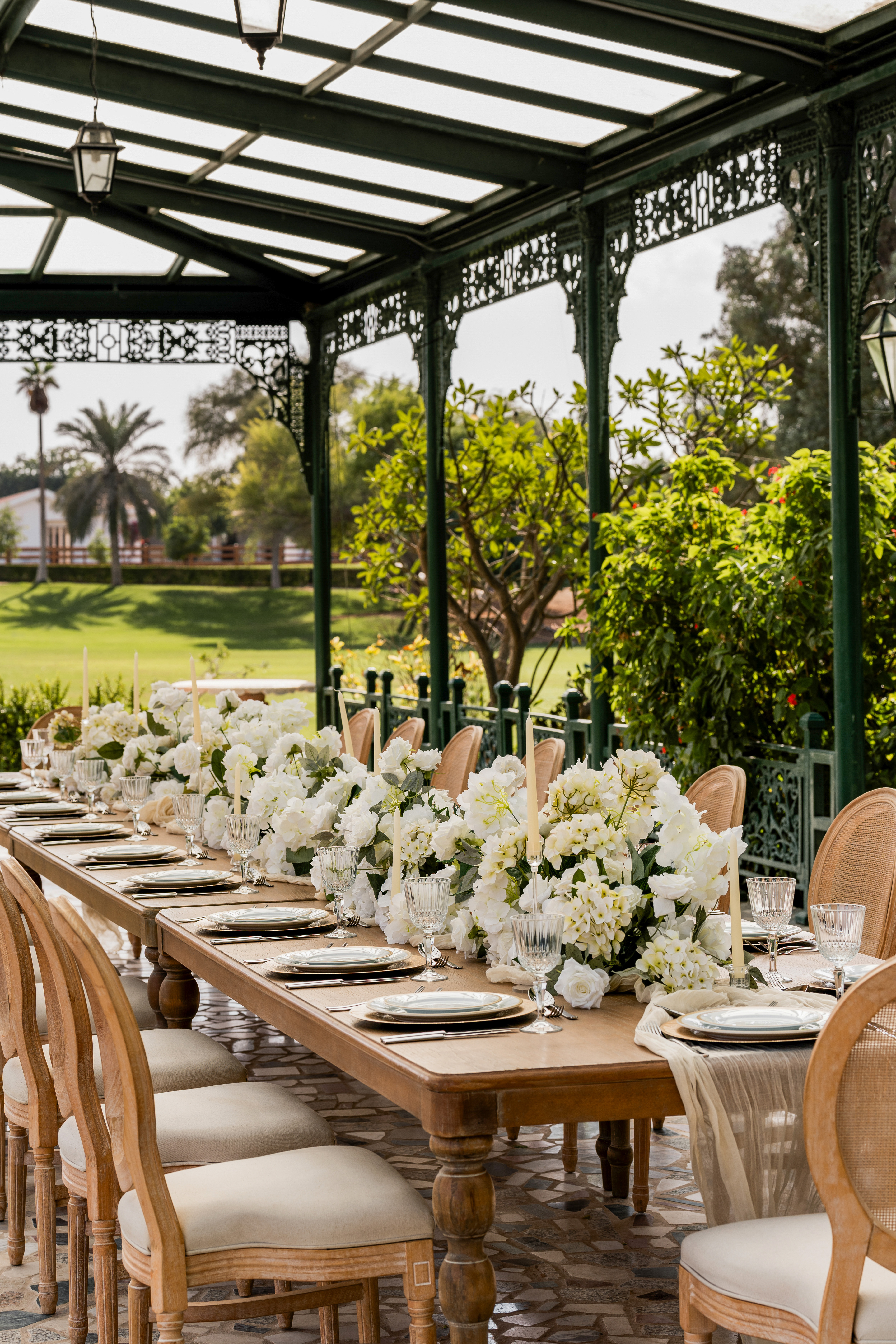 Elegant outdoor dining table with white floral centerpieces under a green gazebo.