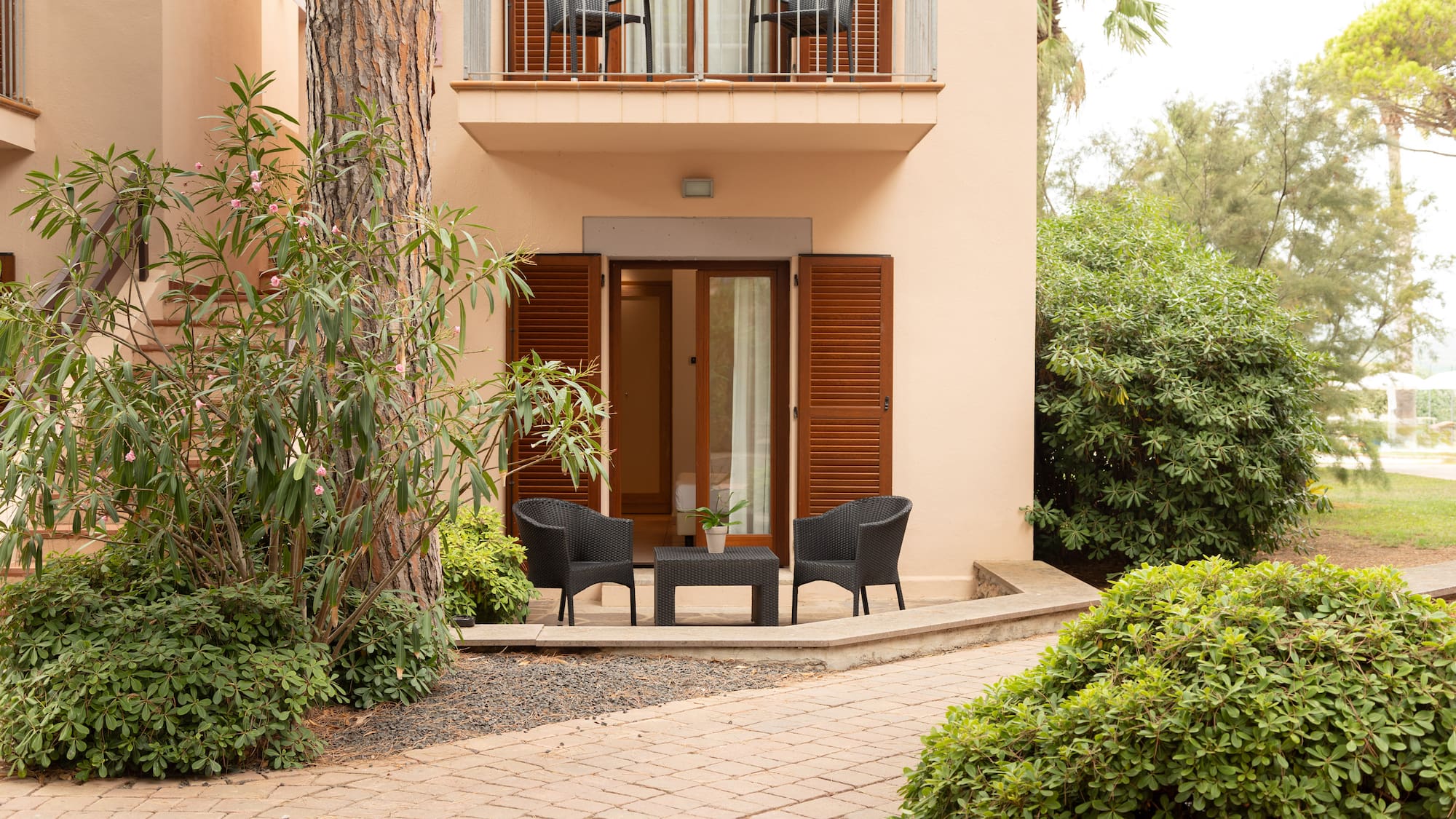 Hotel room patio with black wicker furniture, open shutters, balcony, and lush garden.