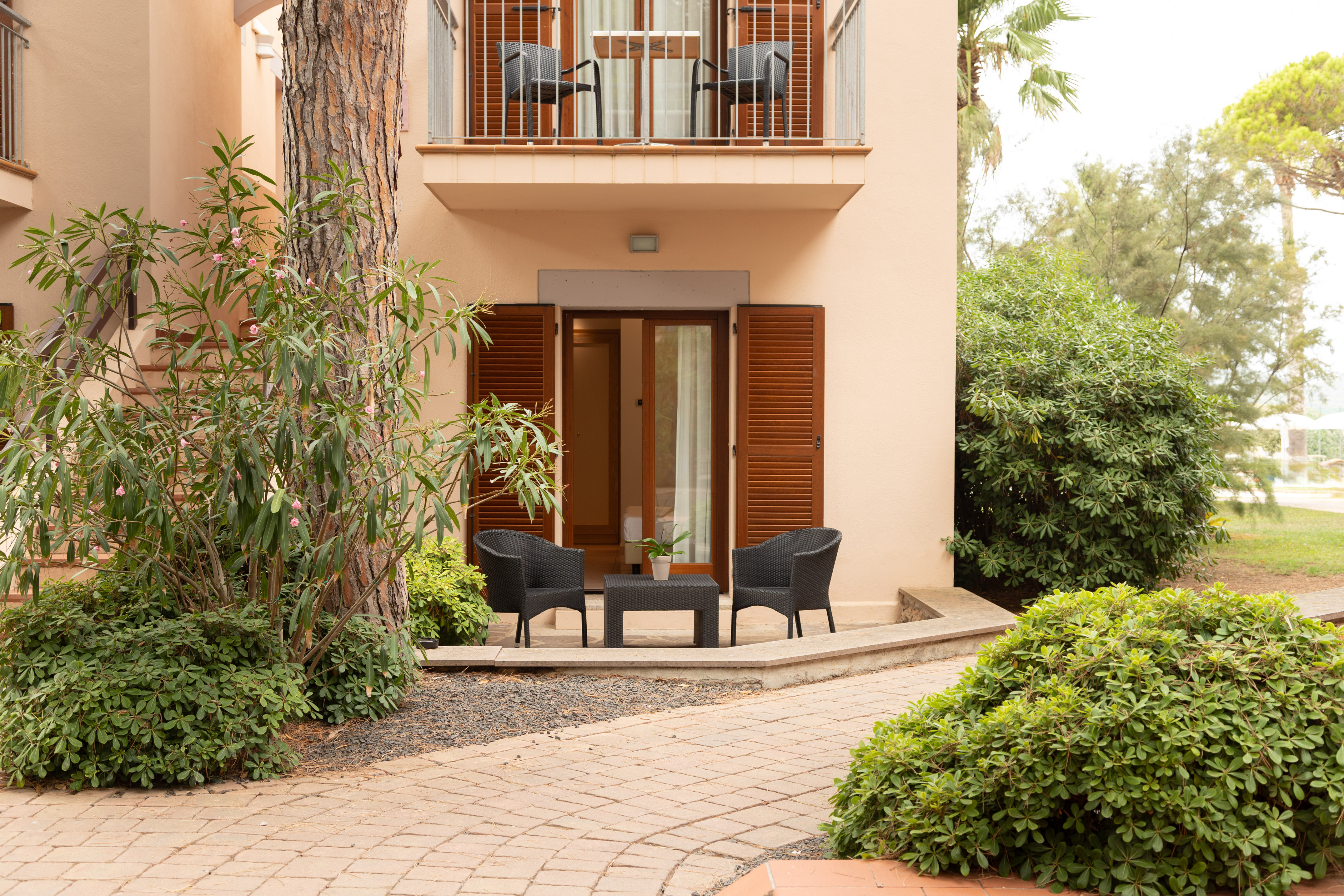 Hotel room patio with black wicker furniture, open shutters, balcony, and lush garden.