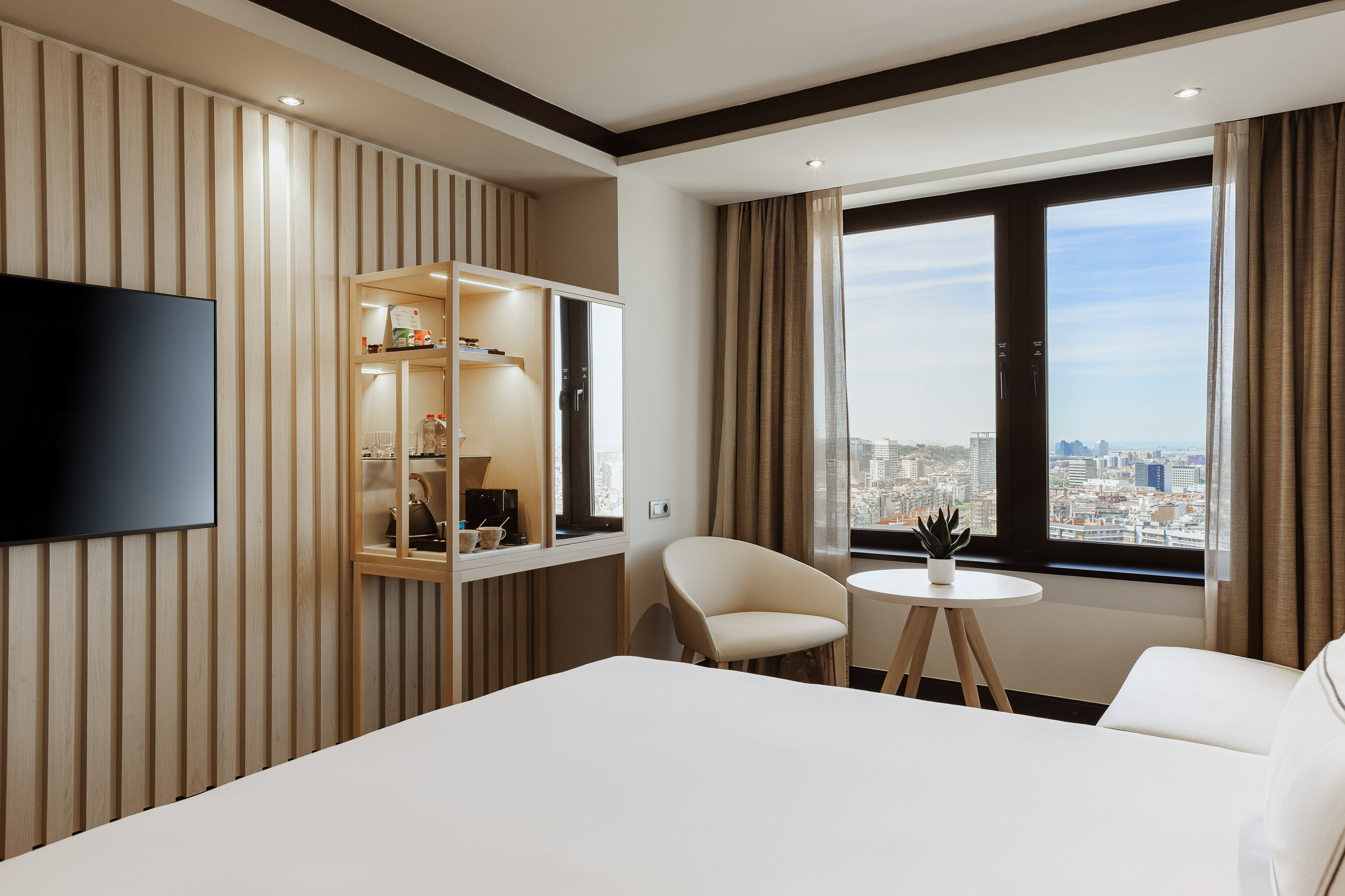 Sleek hotel room with bed, TV, wooden paneling, mini-bar, and city view.