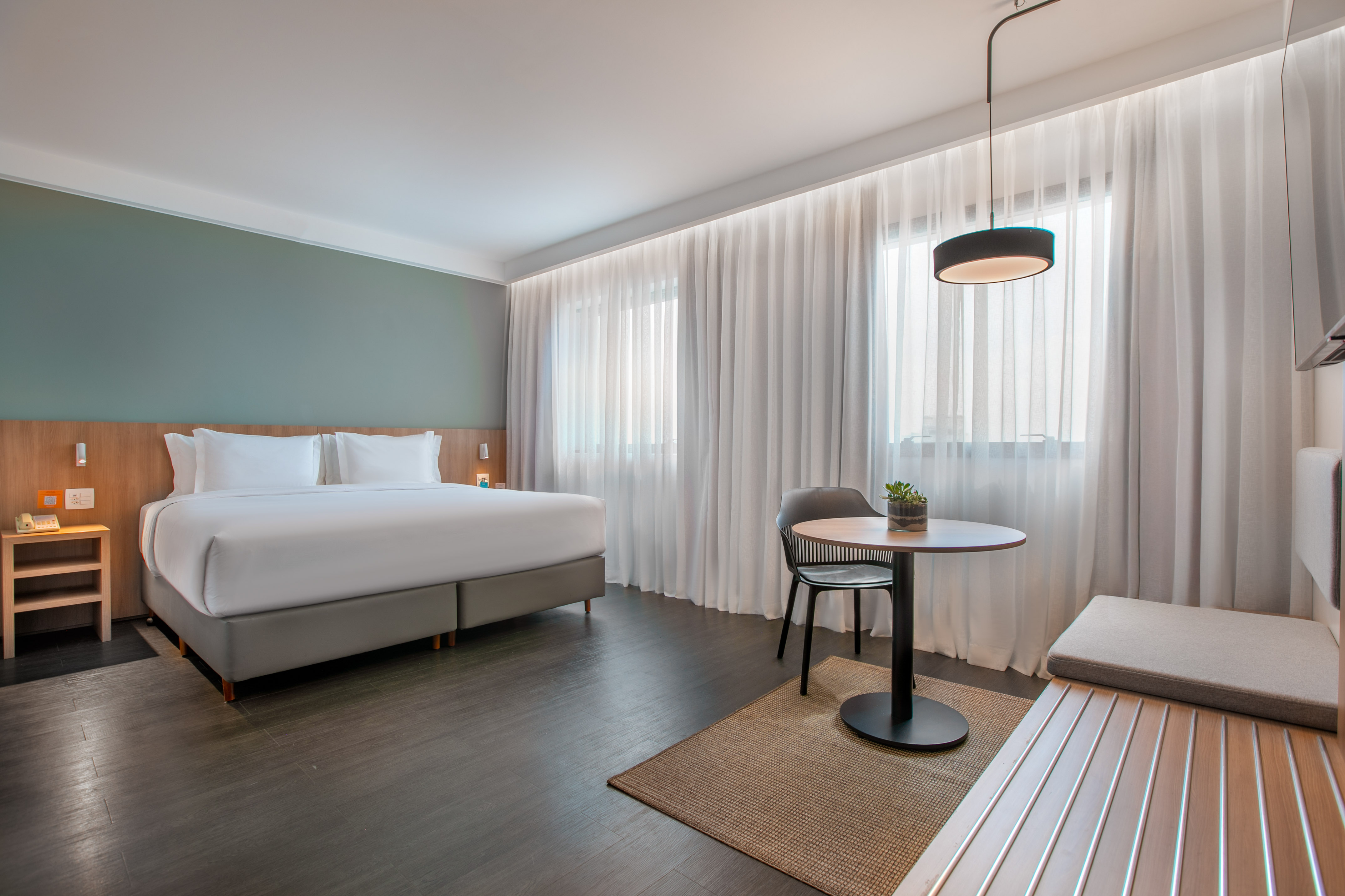 Stylish hotel room with king bed, wood floor, small table, bench, and window.