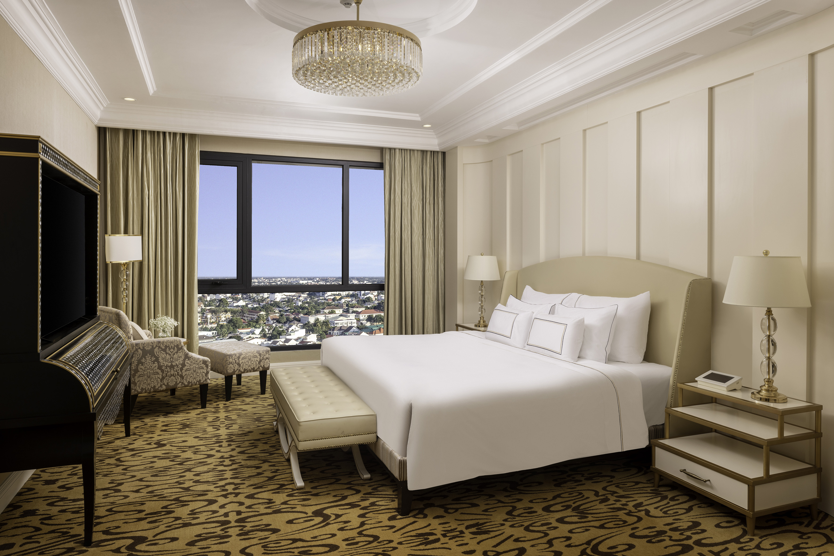 Elegant hotel bedroom with king bed, city view, chandelier, and patterned carpet.