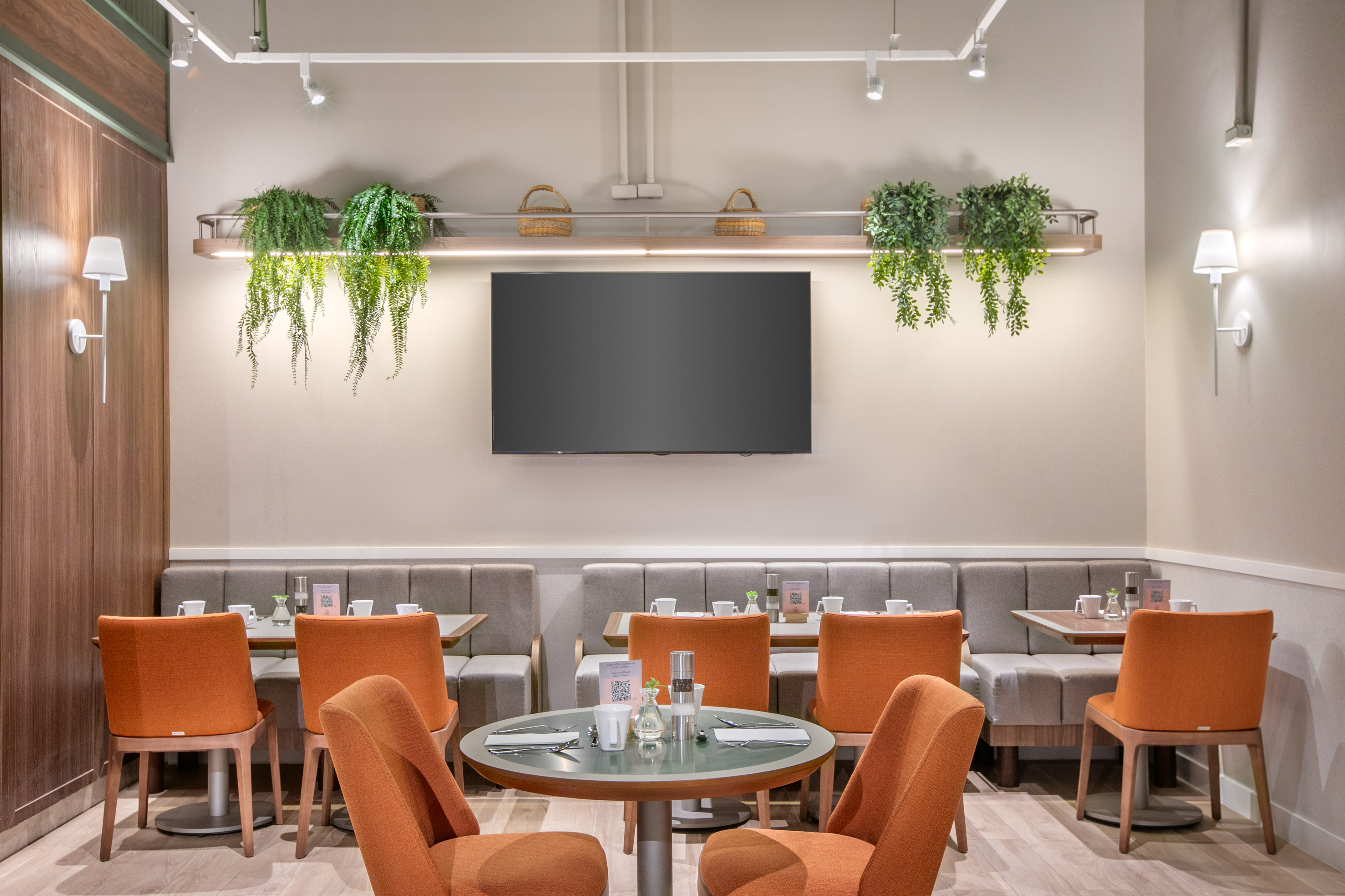 Modern restaurant interior with orange chairs, grey booths, wall TV, and plants.