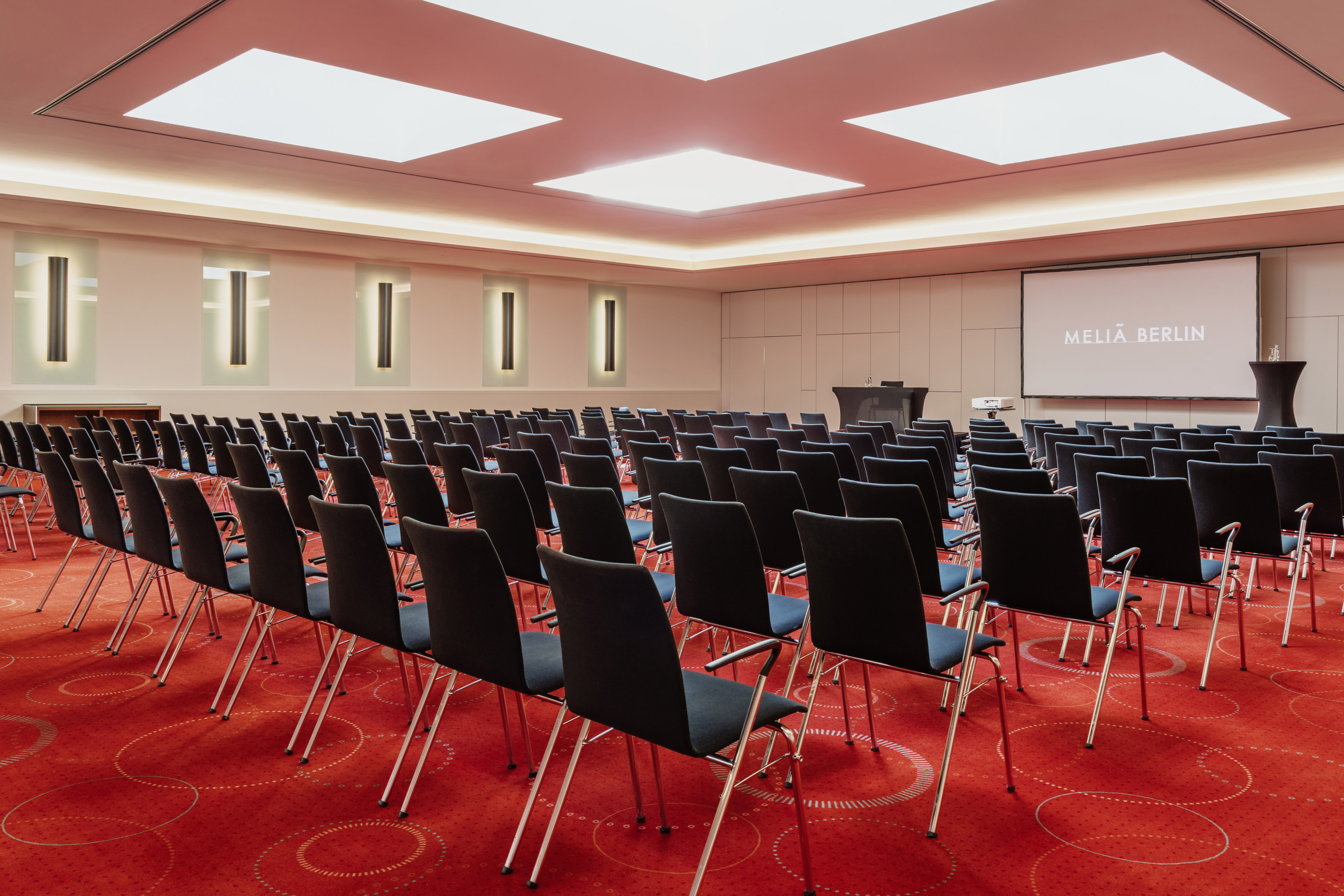Meliá Berlin meeting room with black chairs, red carpet, and projector screen. - Barcelona 1-3 - Meliá Berlin, Berlín