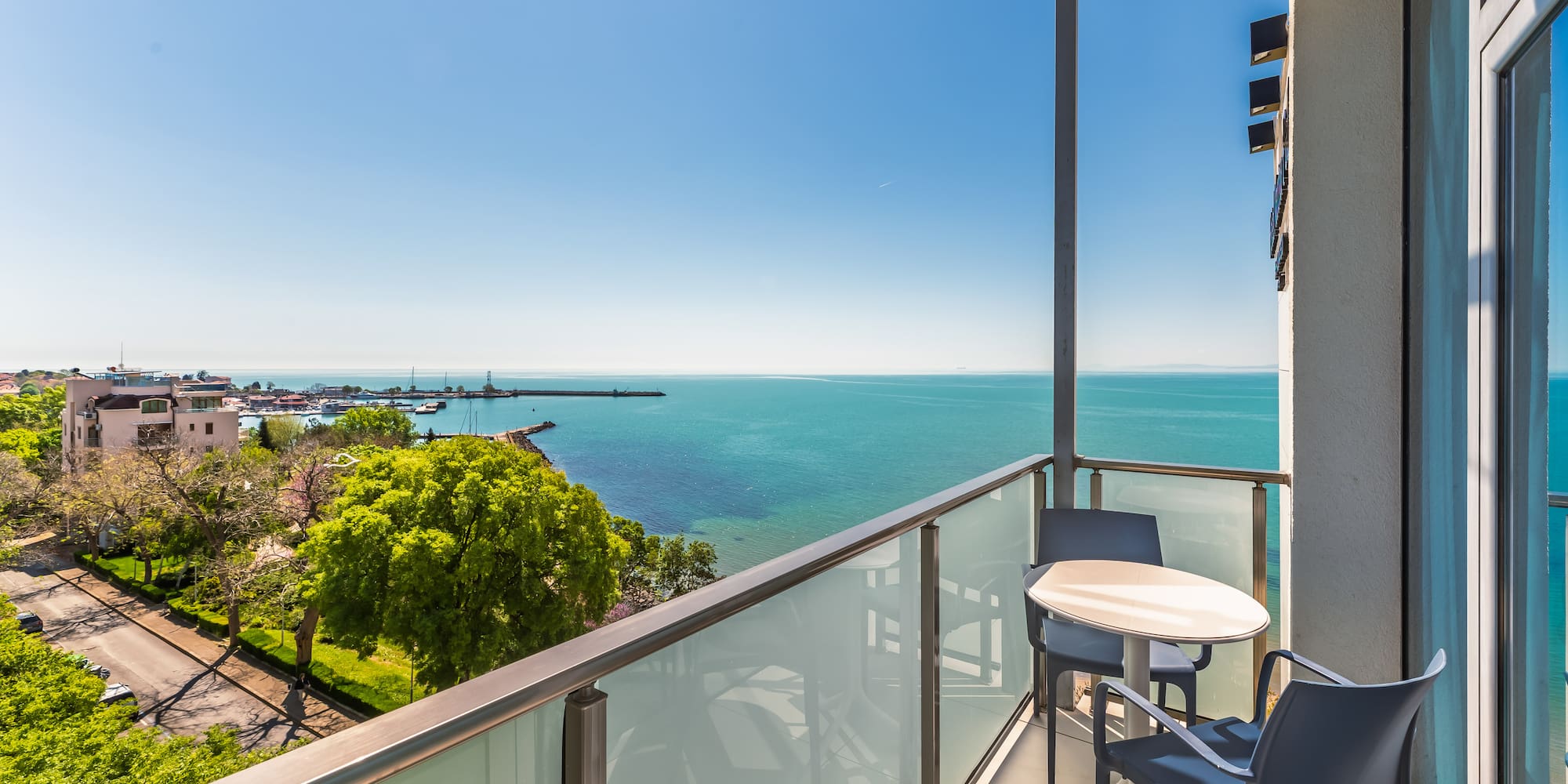 Sunny balcony overlooking turquoise sea and coastal marina with boats.