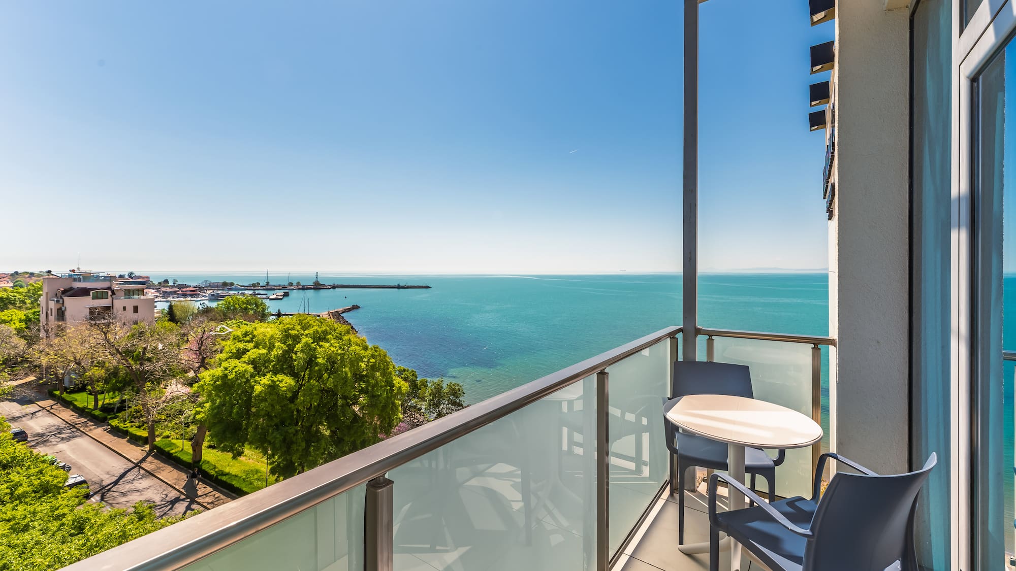 Sunny balcony overlooking turquoise sea and coastal marina with boats.