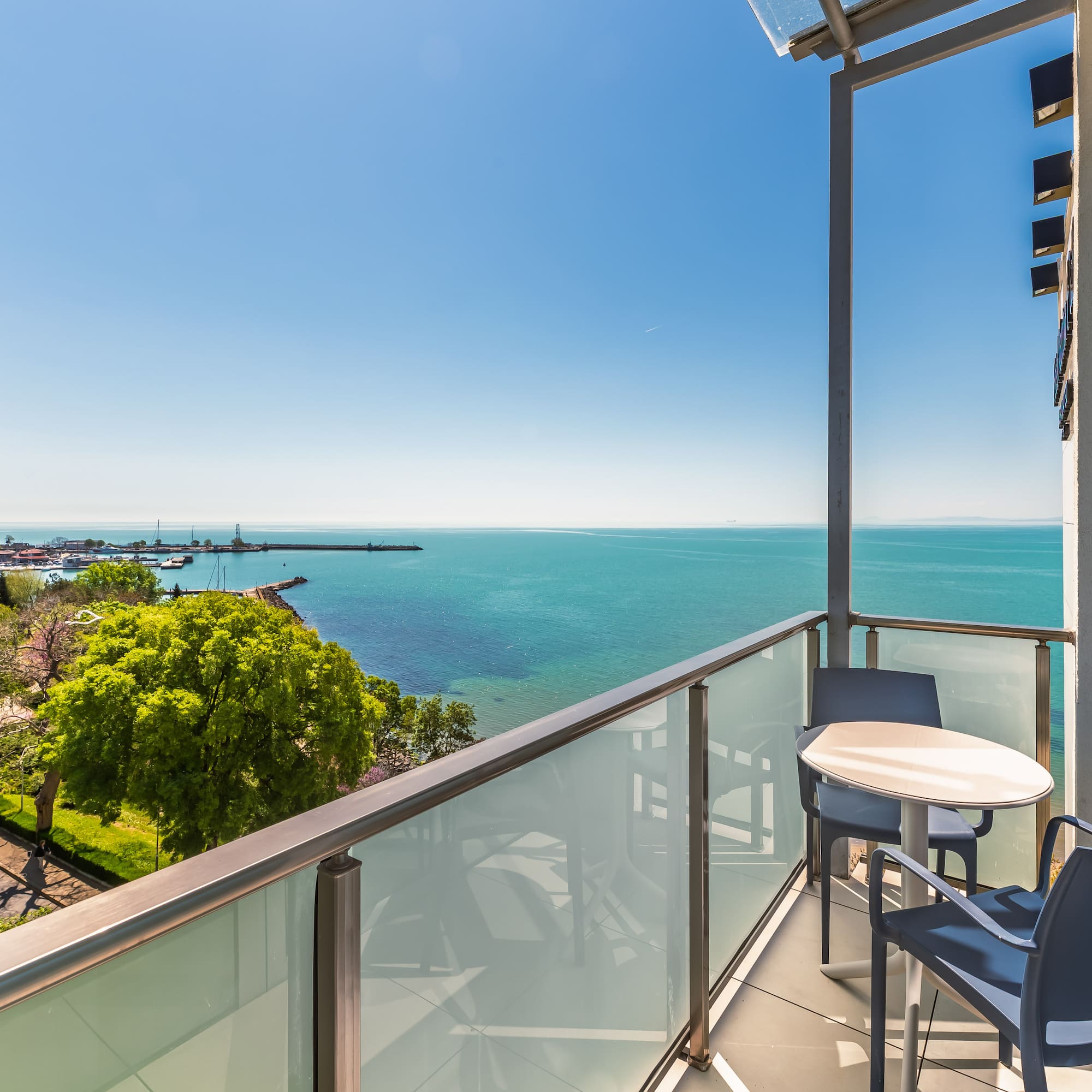 Sunny balcony overlooking turquoise sea and coastal marina with boats.